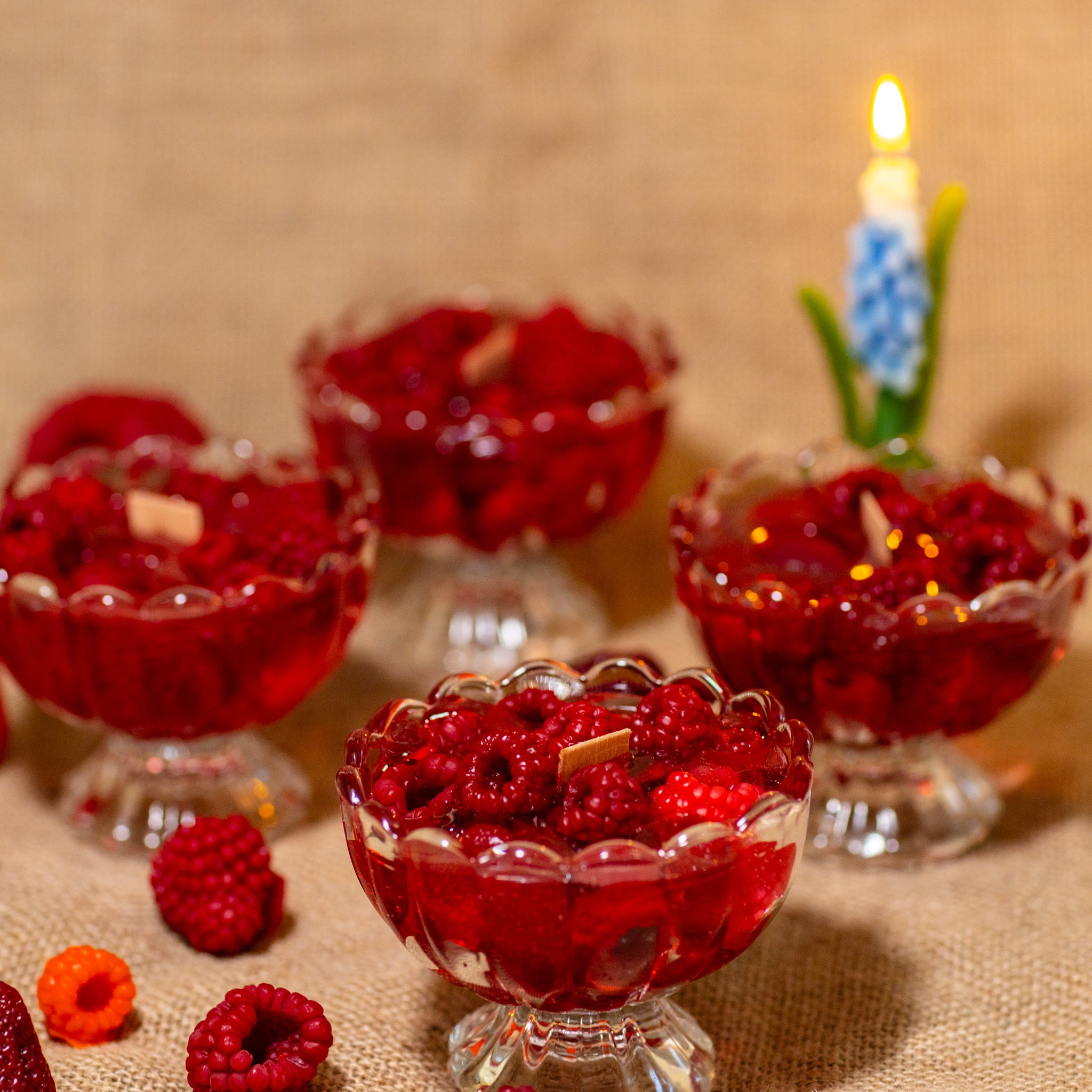 Group of artisan raspberry jam candles in glass dessert dishes arranged on a rustic jute fabric surface, one with a burning candle in the background.
