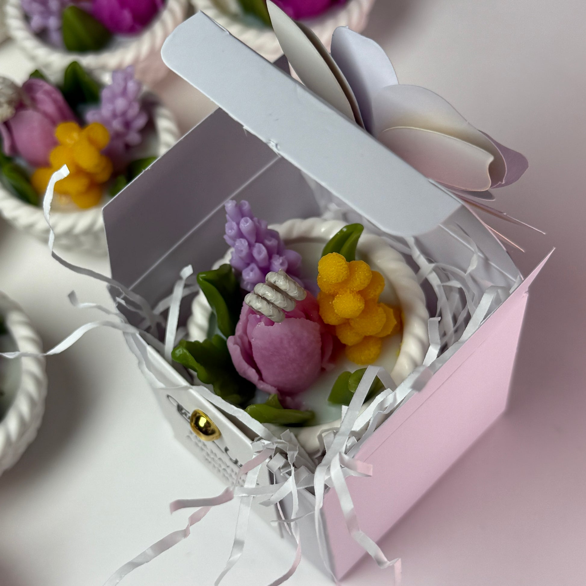 An overhead view of a miniature Jesmonite basket candle neatly packed inside an opened pastel pink and white favour gift box filled with protective shredded paper.