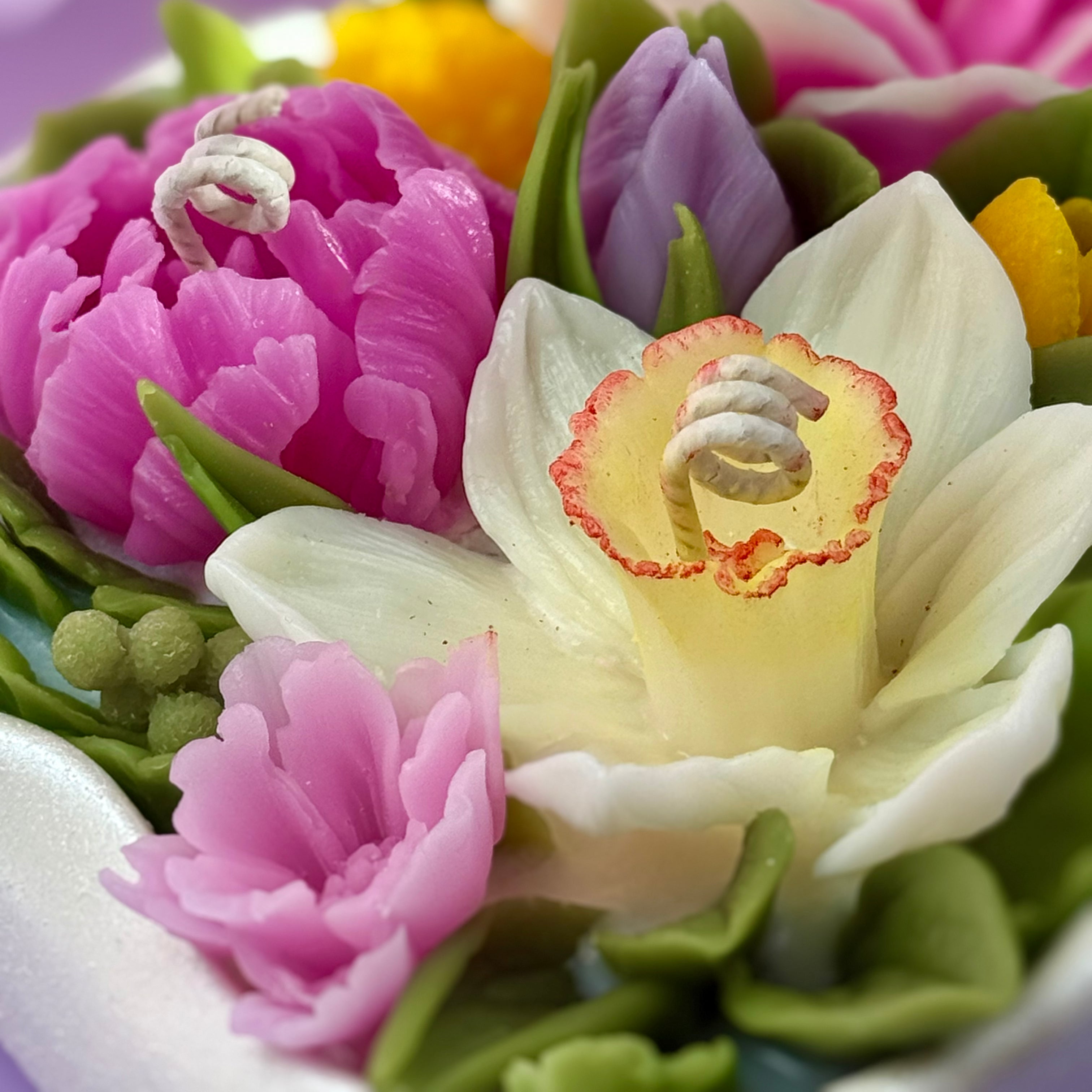 Macro close-up of a hand-sculpted beeswax narcissus and pink peony with detailed petal textures and cotton wicks.