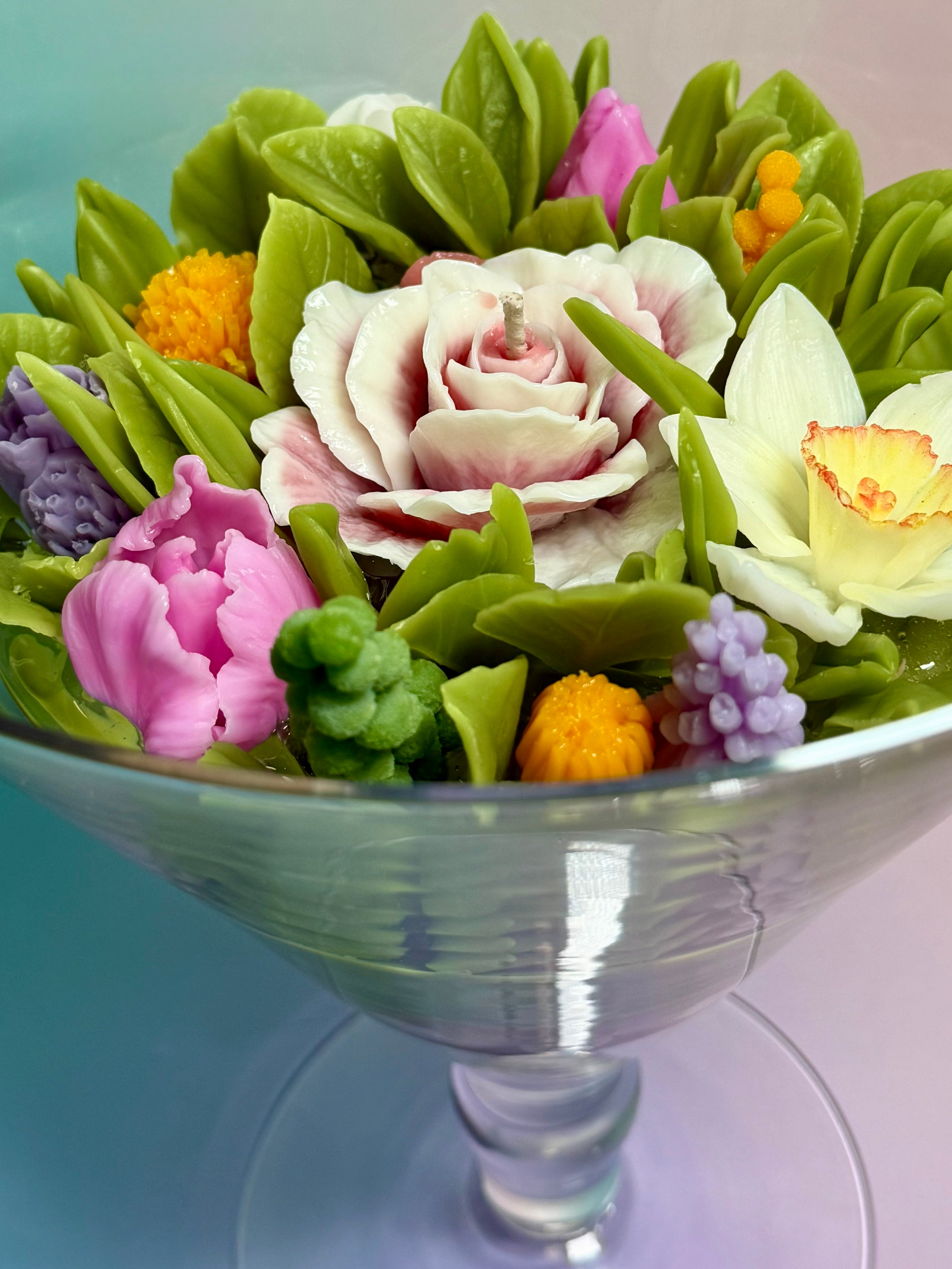 Close-up detail of realistic beeswax flowers and green leaves encased in crystal-clear gel wax within a large glass vase.