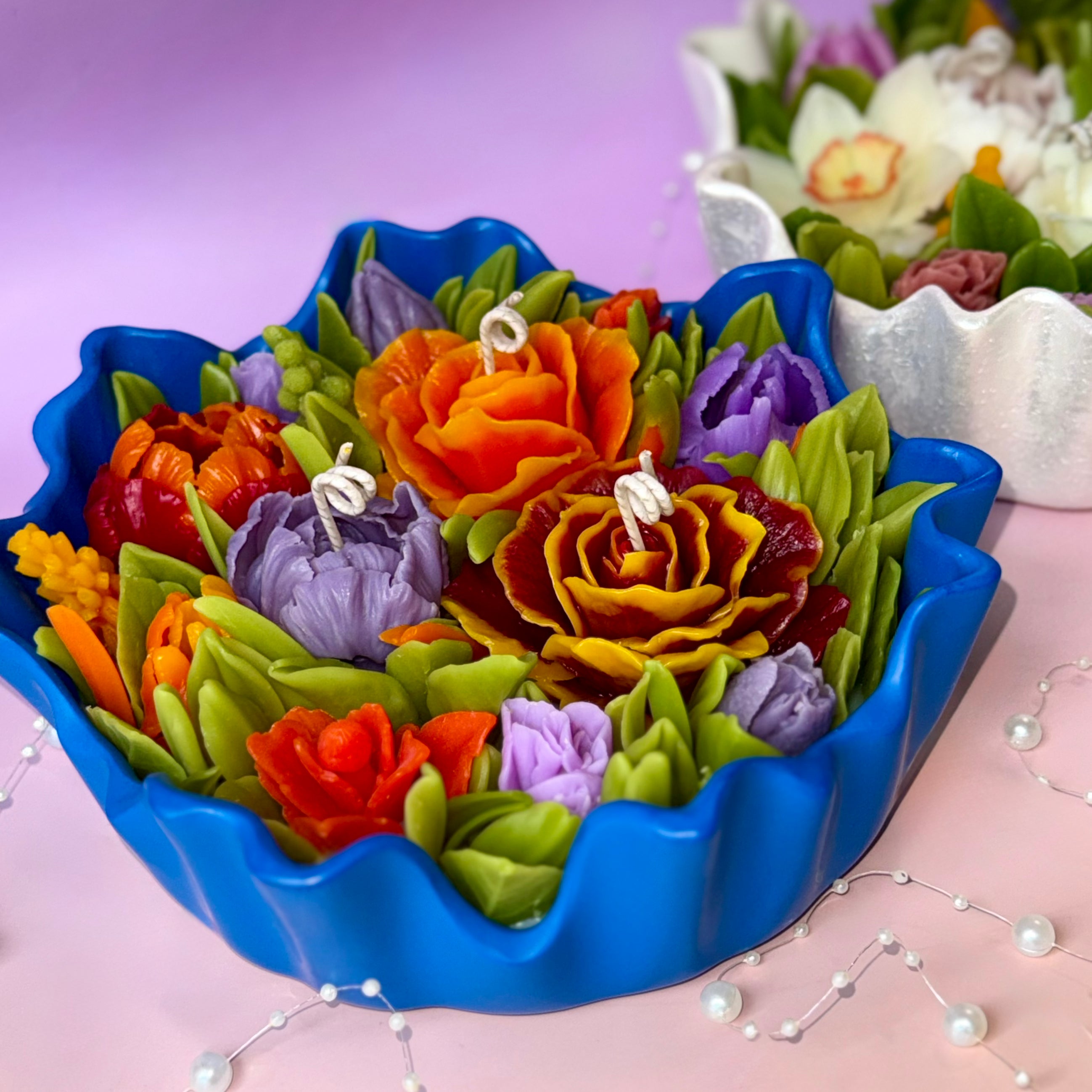 A large 20cm floral centrepiece candle in a vibrant azure blue fluted bowl, featuring a colourful handcrafted beeswax flower bouquet on a pink to purple gradient background with pearl decorations.