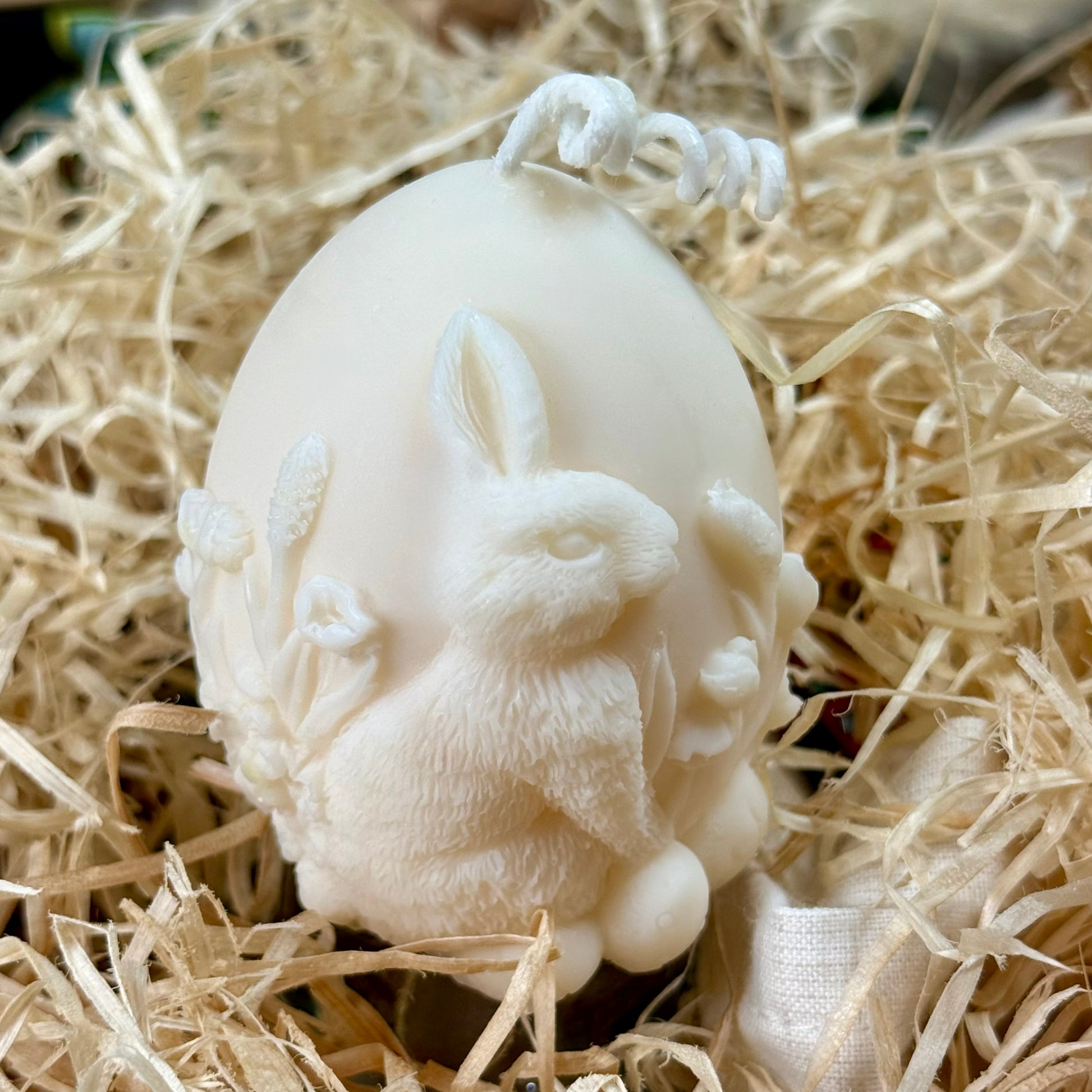 A 115g sculptural Easter bunny egg candle made of cream-coloured natural soy wax, nestled in a bed of decorative straw shredded paper, highlighting the detailed 3D bunny and floral relief.