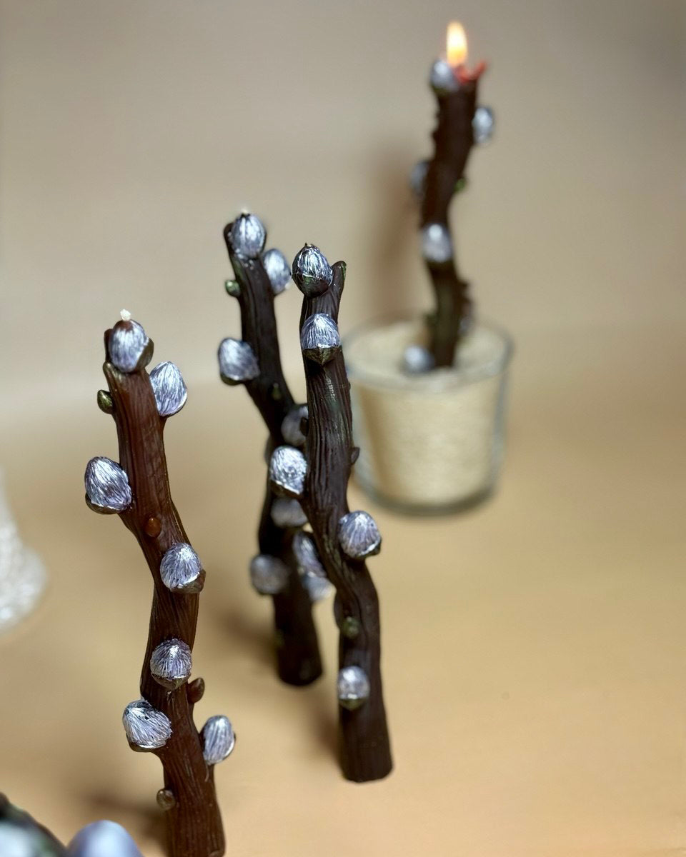 Multiple handmade beeswax willow branch candles arranged together on a warm beige backdrop, with one candle lit and glowing softly in a glass base in the background.