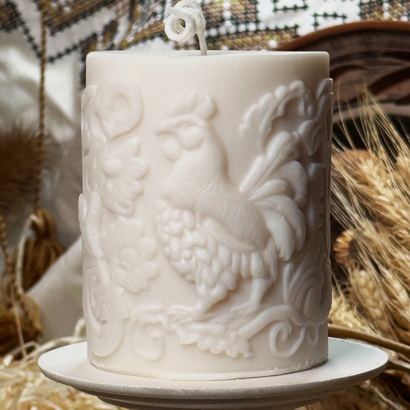 A side-angle shot of a creamy white artisanal botanical rooster pillar candle resting on a small white pedestal dish, with a background of dried wheat stalks and traditional patterned embroidery.