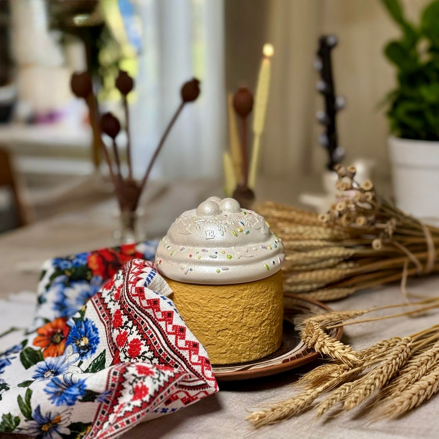 An artisanal Easter Paska candle designed like traditional holiday bread with a textured yellow base and a pearlescent white icing lid, styled on a table with a floral patterned towel and dried wheat ears.