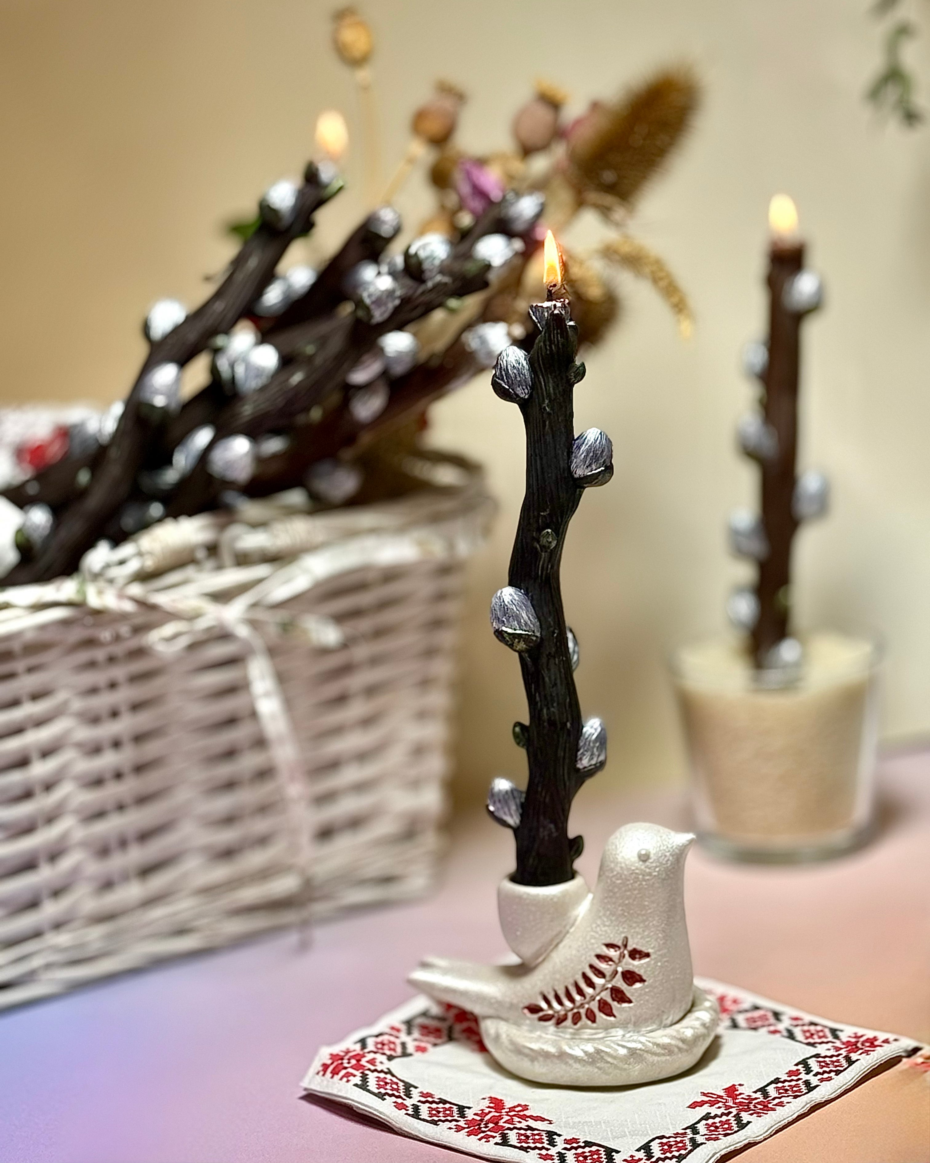 A burning dark beeswax willow branch candle held securely by a pearl-white Jesmonite bird holder with hand-painted red leaves, resting on a traditional embroidered napkin.