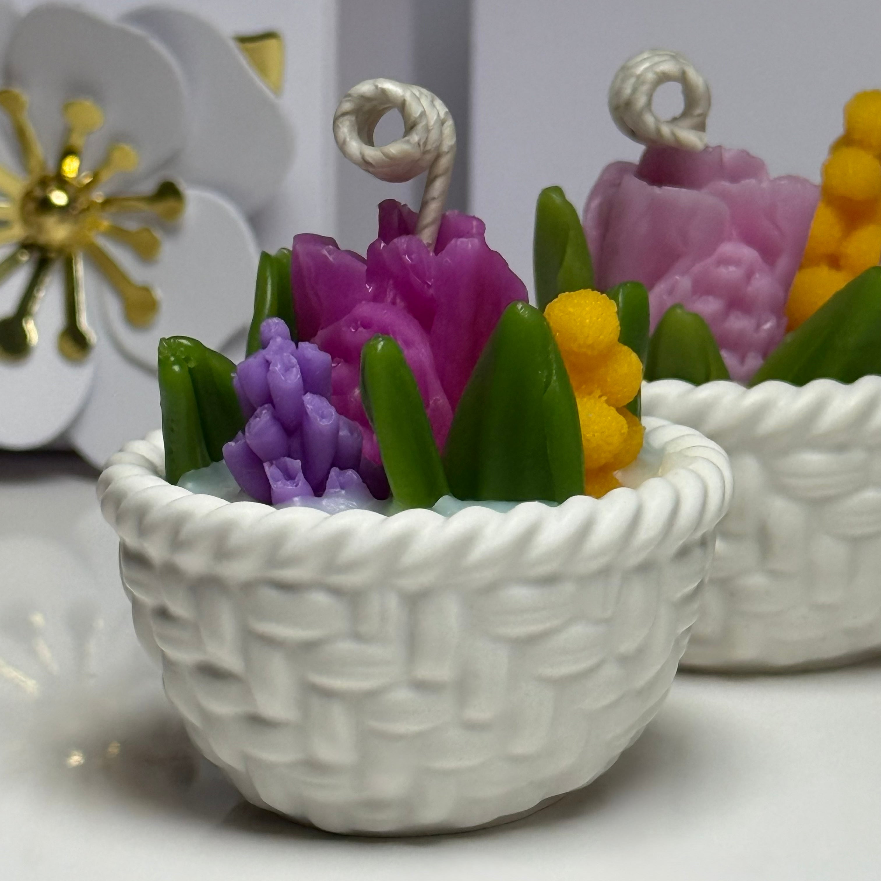 A close-up shot of two hand-cast white Jesmonite basket candles, highlighting the detailed rope-weave design filled with vibrant purple, pink, and yellow hand-sculpted beeswax spring flowers.