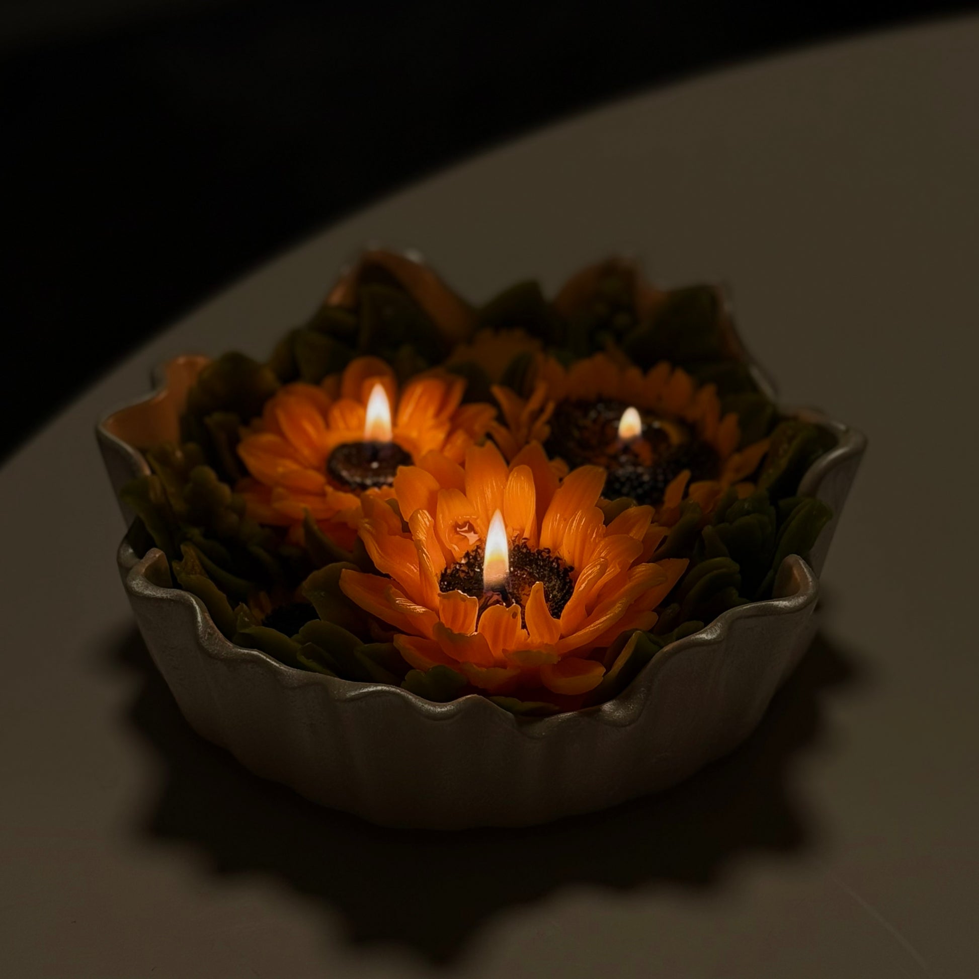 Cosy atmospheric shot of a large burning sunflower multi-wick statement candle with three glowing wicks inside a hand-painted pearl white Jesmonite scalloped bowl.