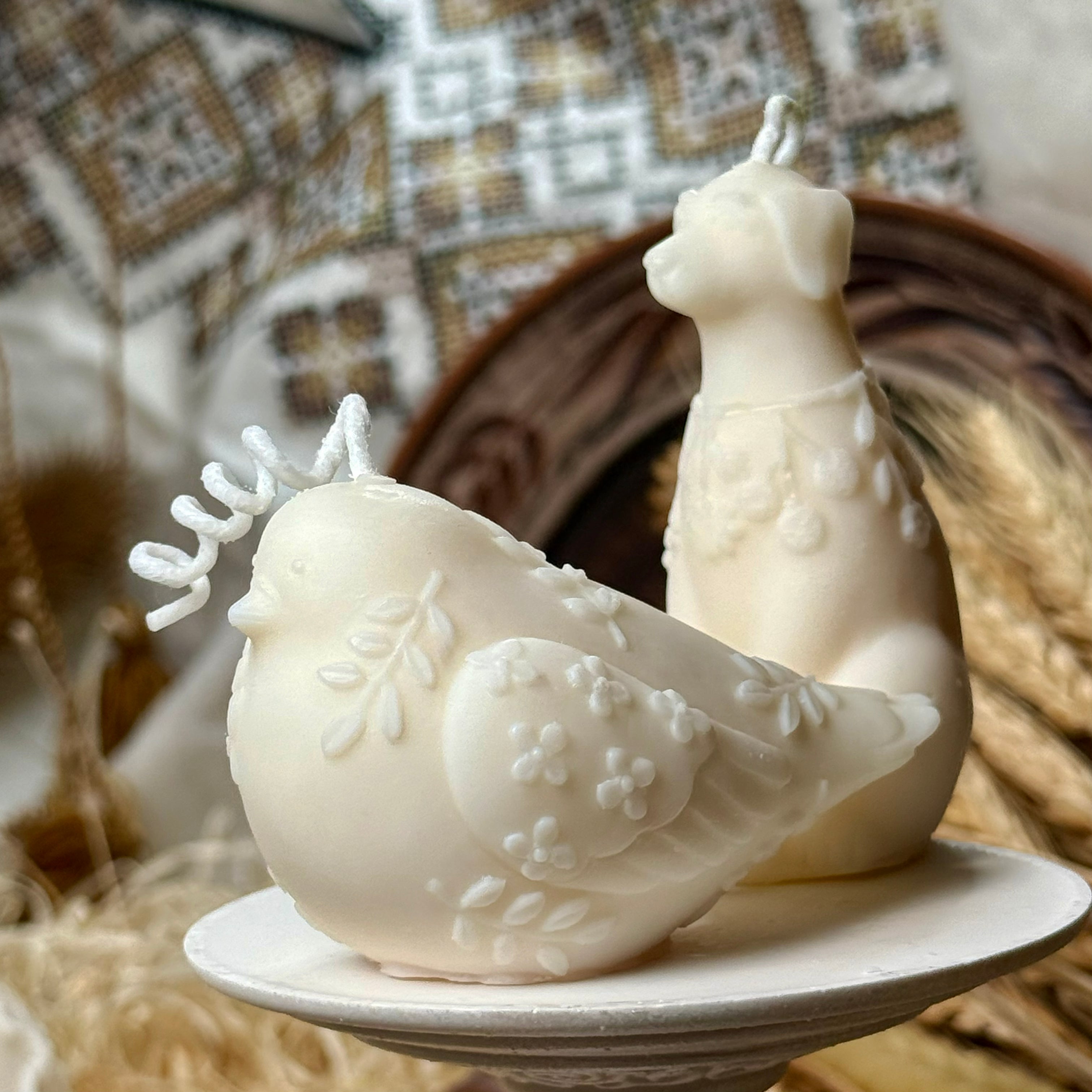 An elegant side profile of the botanical bird candle with detailed leaf patterns on its wings, sitting on a white pedestal next to the matching botanical dog candle.
