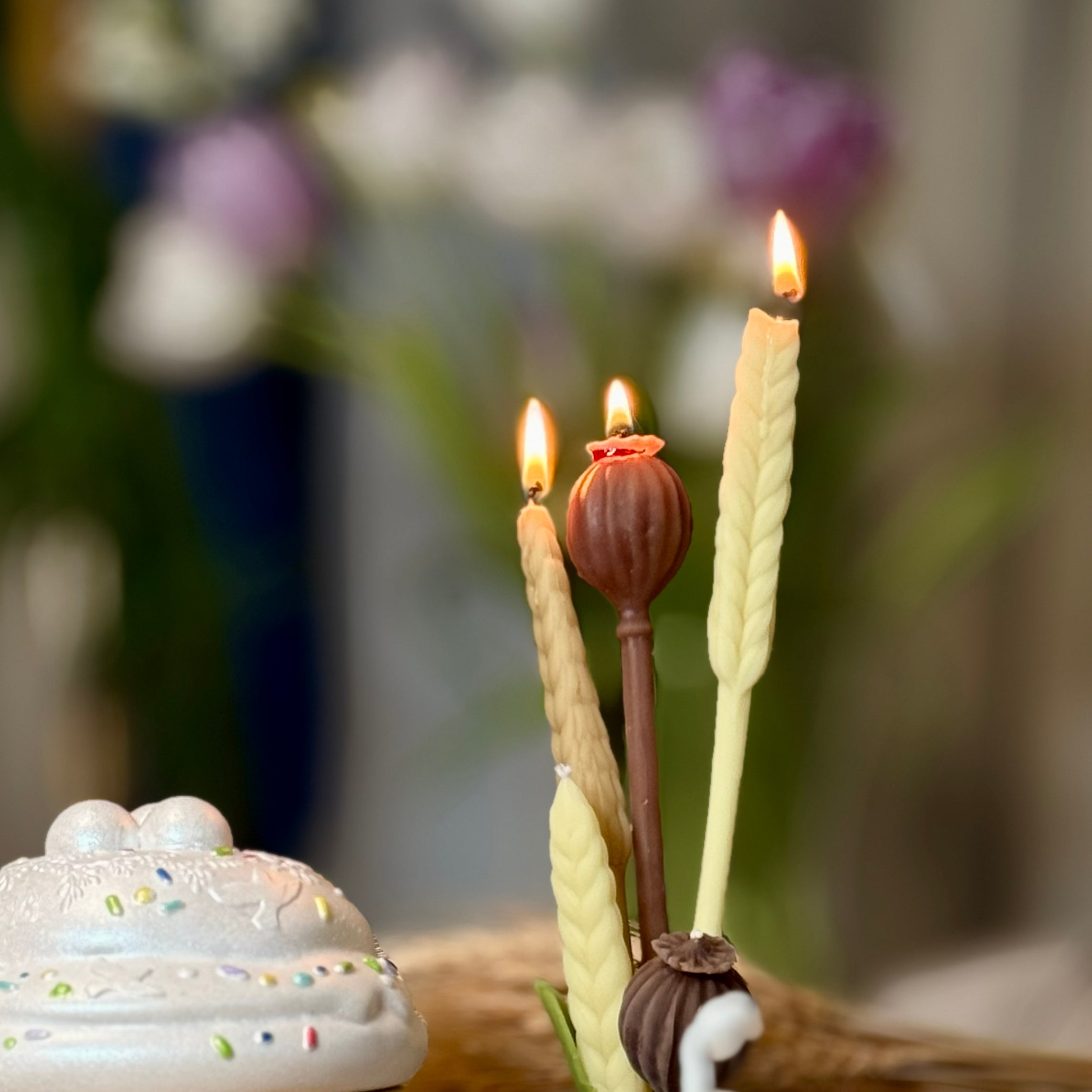 A side-by-side shot of the artisanal botanical pure beeswax candles, featuring the detailed golden ivory Wheat Stalk taper and the earthy ochre Poppy Pod candle sitting on a neutral linen cloth.