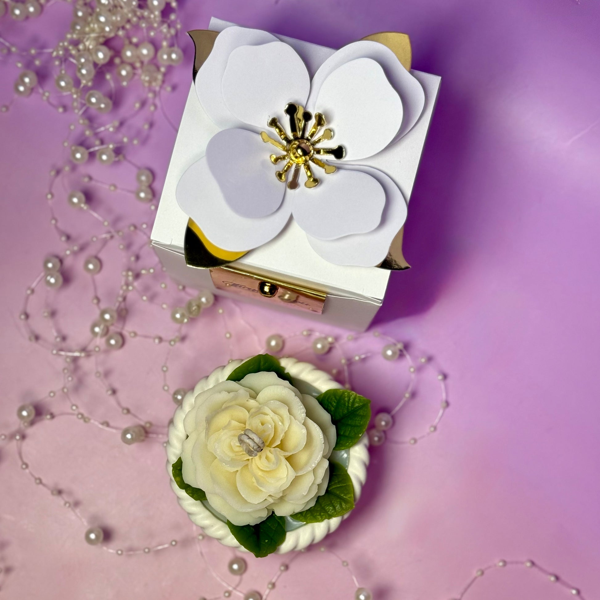 High-angle view of the white cream rose basket candle sitting next to its premium boutique box which is decorated with a luxury white paper flower structure.