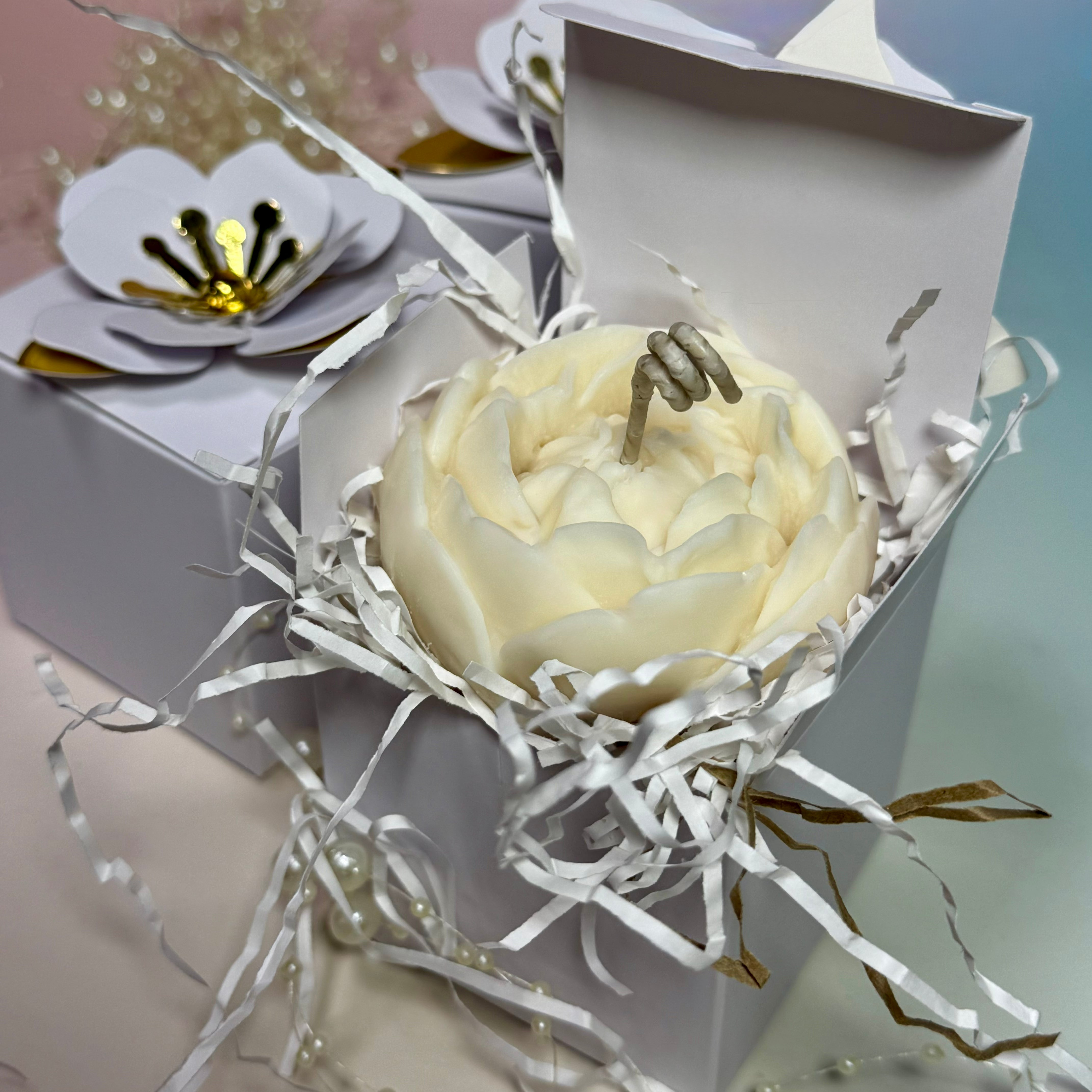 High-angle shot of a white gift box with an artistic 3D white paper magnolia topper and gold leaves, partially open showing a handcrafted peony flower candle inside.