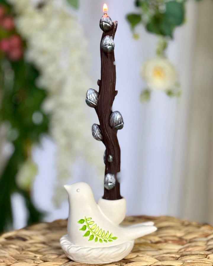 A textured brown beeswax pussy willow candle standing tall in a white Jesmonite bird candle holder decorated with vibrant hand-painted green leaves on a rustic wicker surface.