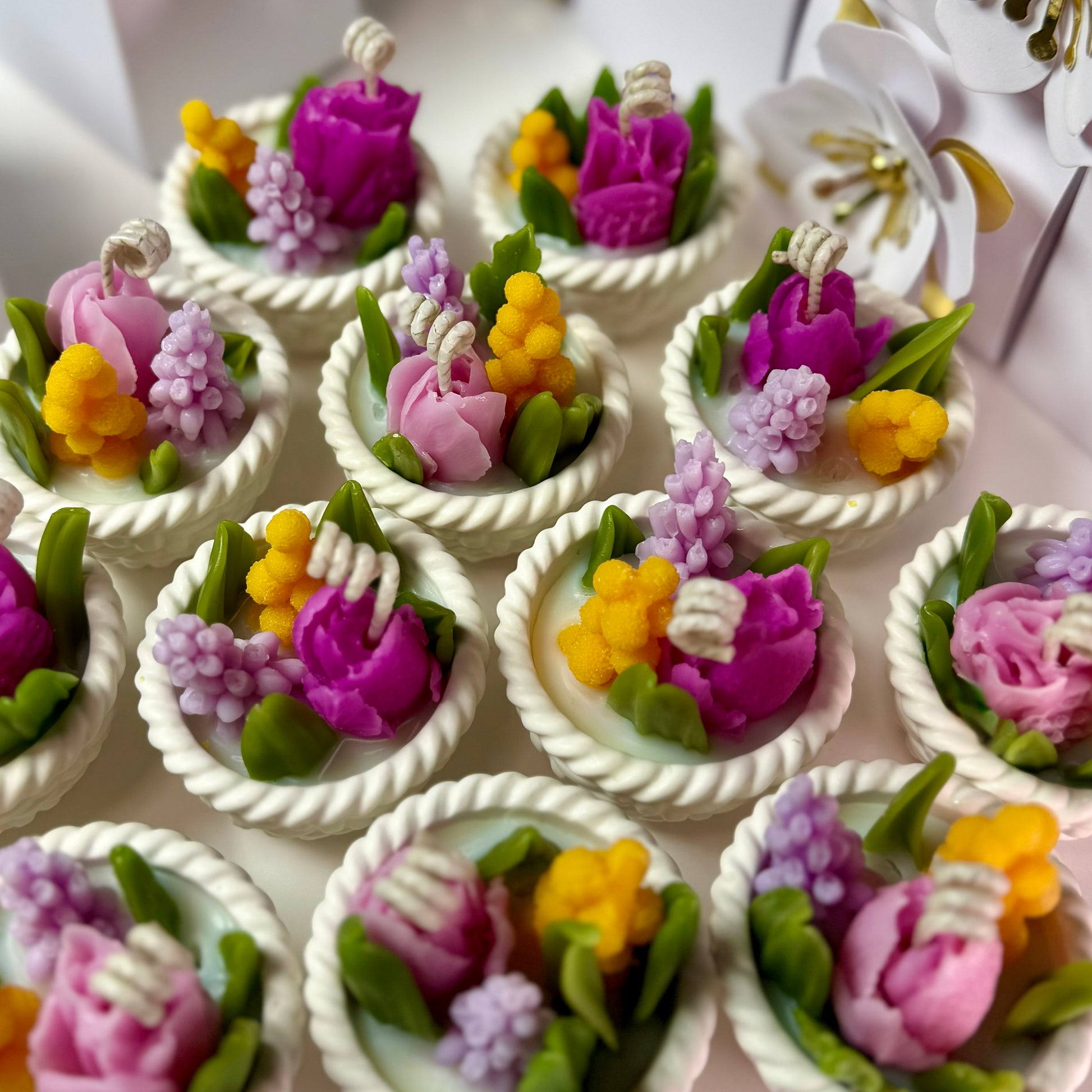 An overhead group shot of multiple mini woven Jesmonite baskets holding vibrant hand-poured soy candles topped with highly detailed pink, purple, and yellow beeswax blossoms.