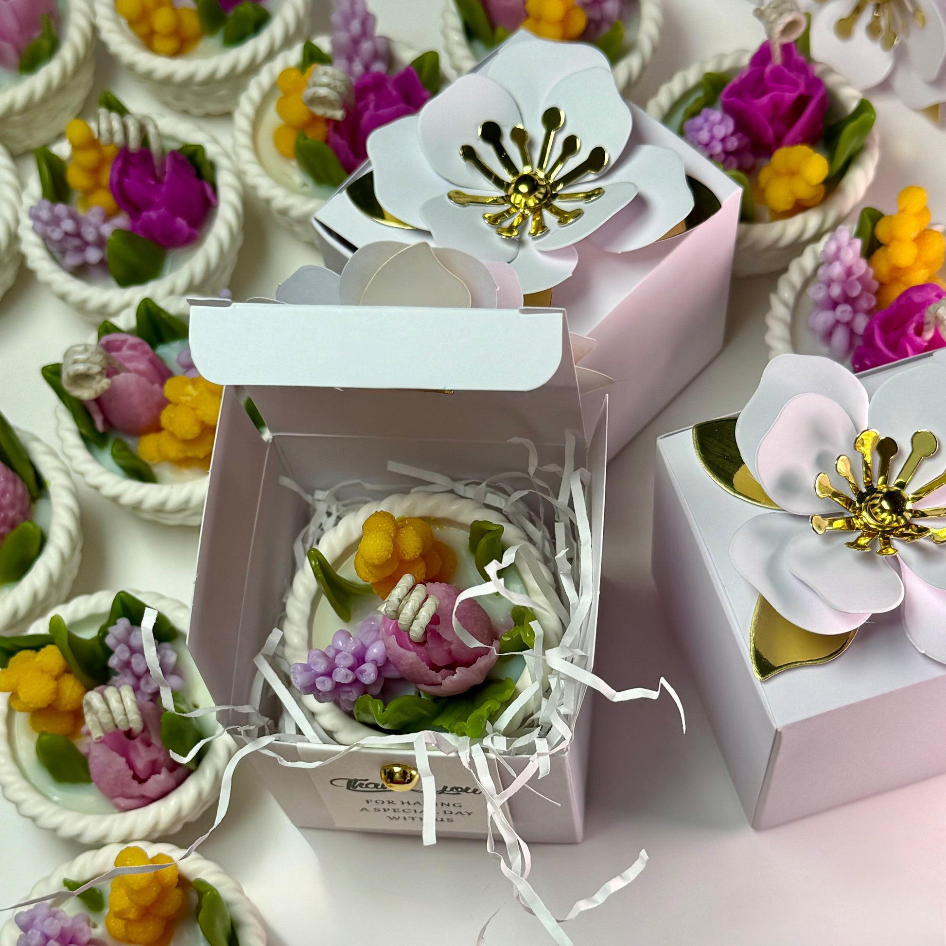 A group display of artisan wedding favour boxes decorated with large luxury white paper flowers, showing one open box to display the botanical soy candle inside.