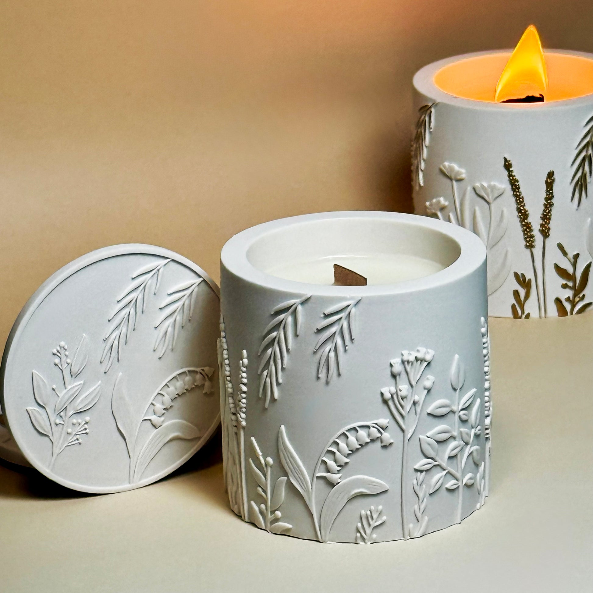 An open classic white botanical soy candle with a wooden wick and its lid resting beside it, styled in front of a burning hand-painted gold botanical relief candle with a bright flame.