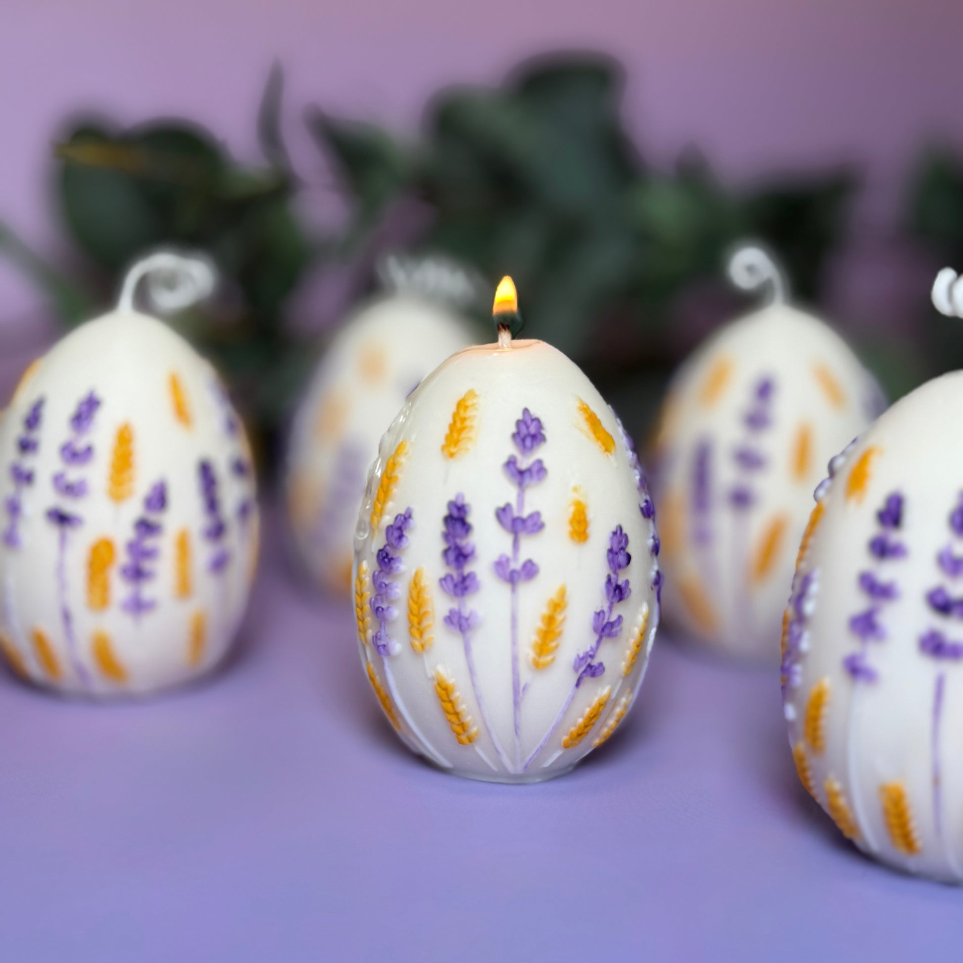 A group of hand-painted botanical egg candles on a purple surface, with the central egg candle lit and casting a warm, gentle flame from its twisted white cotton wick.