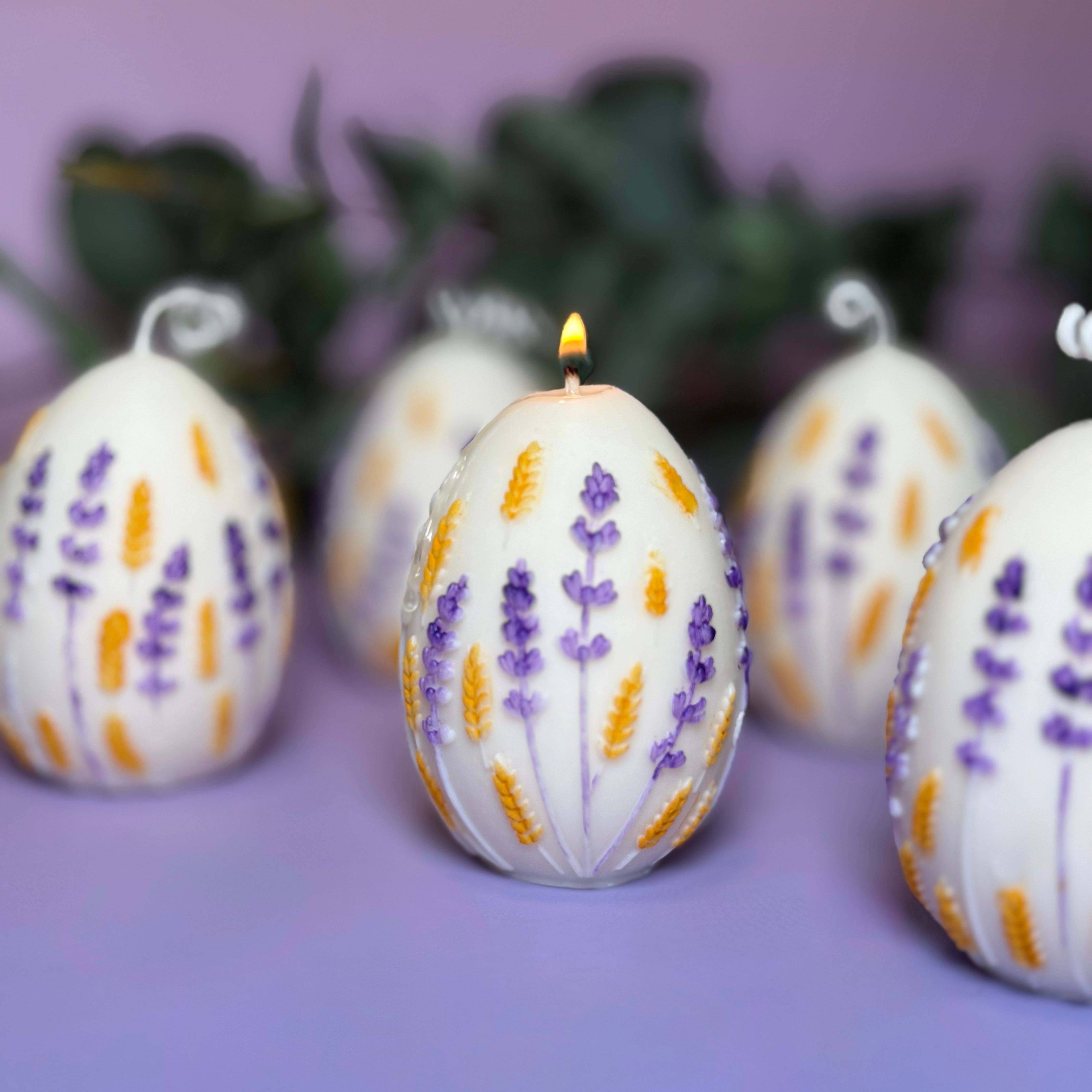 A group of hand-painted botanical egg candles on a purple surface, with the central egg candle lit and casting a warm, gentle flame from its twisted white cotton wick.