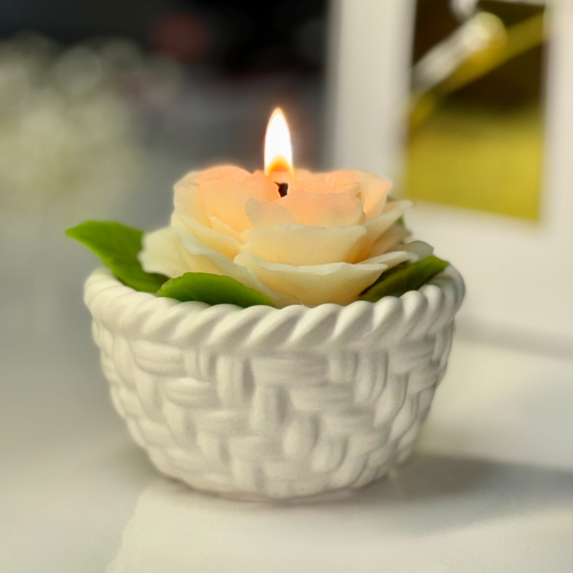 A close-up view of a lit creamy white beeswax rose candle in its woven basket base, showing a warm glowing amber flame casting light on the delicate wax petals.