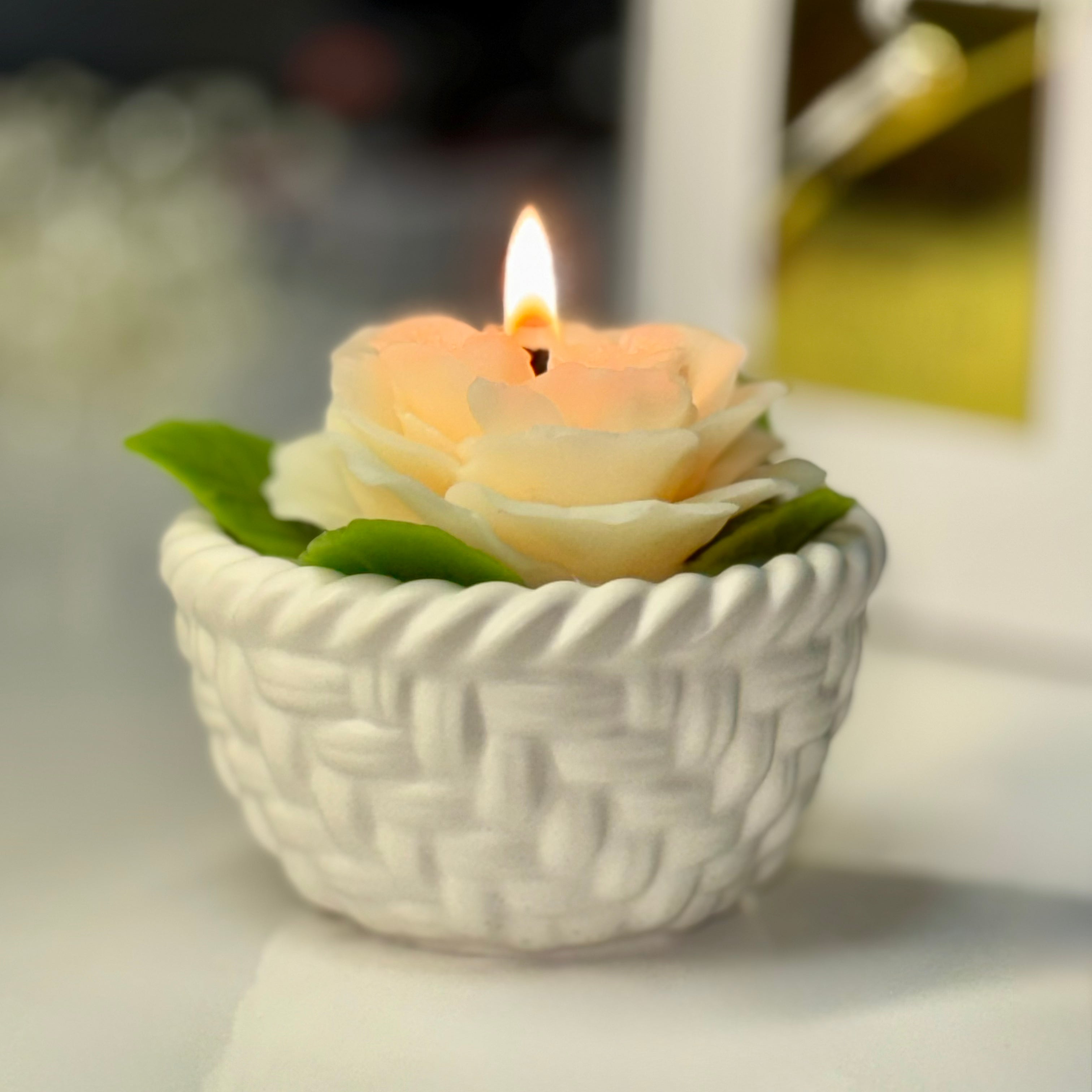 A close-up view of a lit creamy white beeswax rose candle in its woven basket base, showing a warm glowing amber flame casting light on the delicate wax petals.