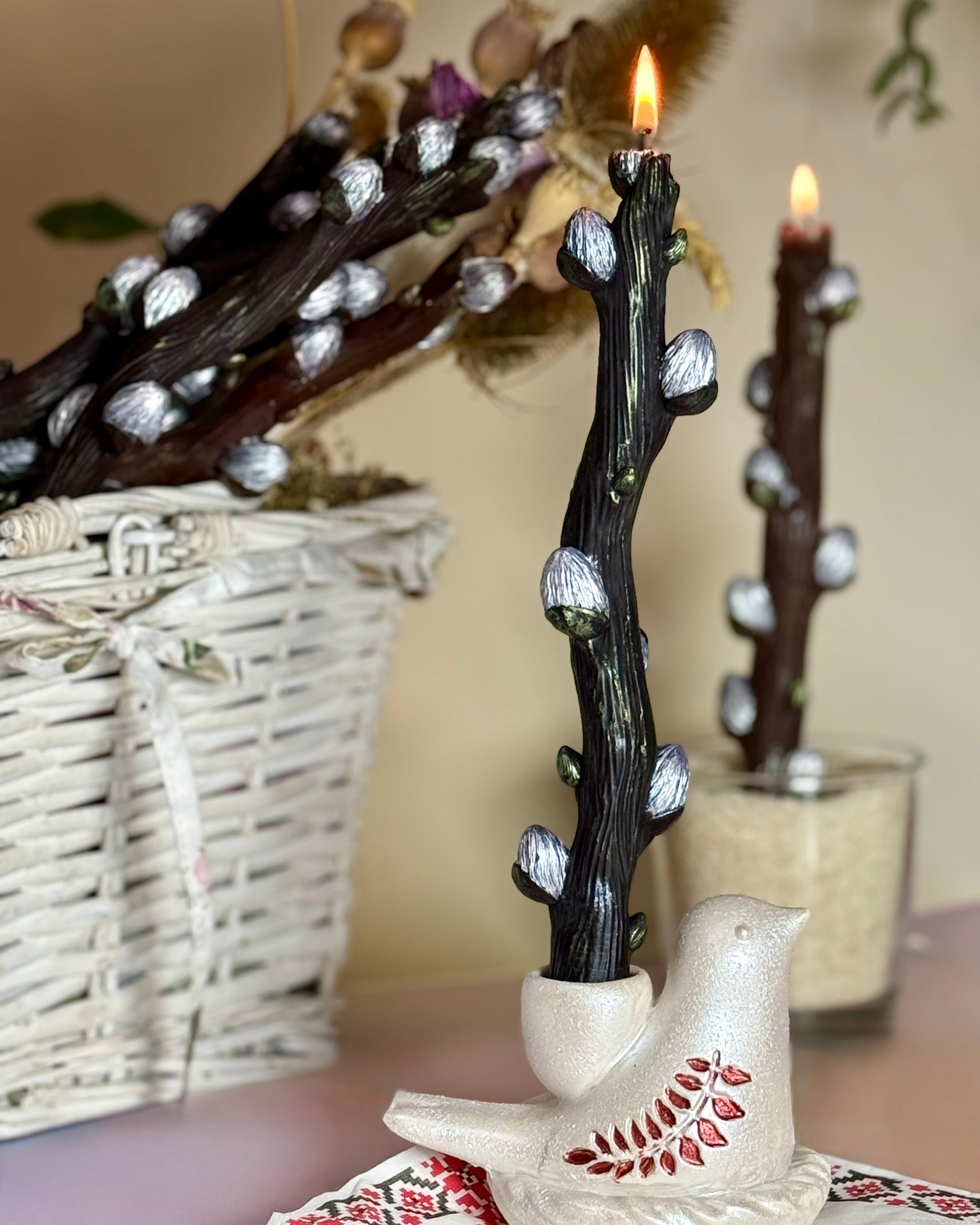 Close-up of a lit black beeswax willow branch candle with silver buds in a hand-painted red leaf Jesmonite bird base, with a basket of willow branches blurred in the background.