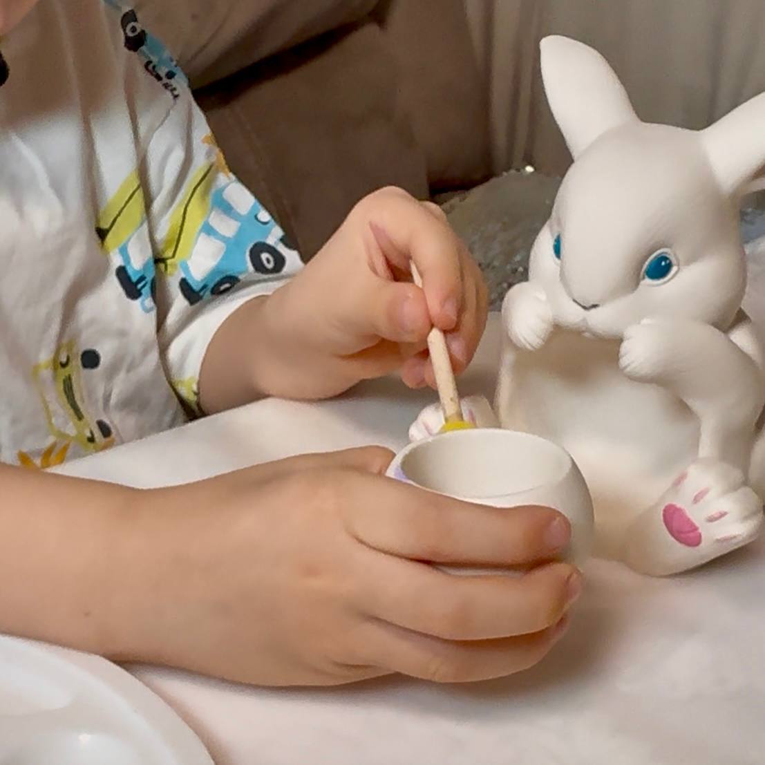 A close-up of a child's hands carefully positioning a cotton candle wick inside the integrated vessel of a hand-cast Jesmonite bunny, with soy wax flakes and paint pots visible in the background as part of a DIY candle making kit activity.