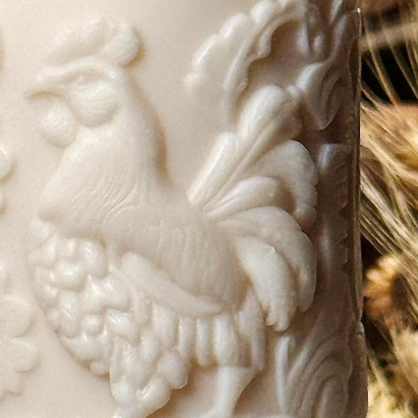 A detailed macro close-up of the sculptural candle's surface, highlighting the crisp and highly intricate relief patterns of the rooster feathers and surrounding folk-art botanical swirls.