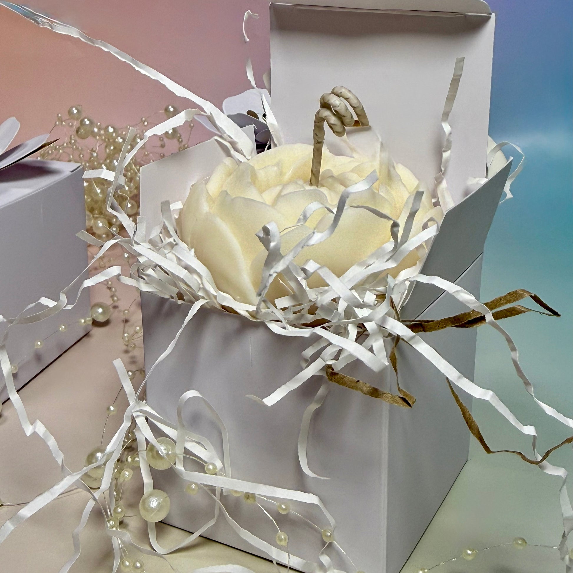 A detailed macro shot into an opened premium white gift box showing a beautiful ivory soy wax peony candle with a twisted wick sitting on white shred filler paper.