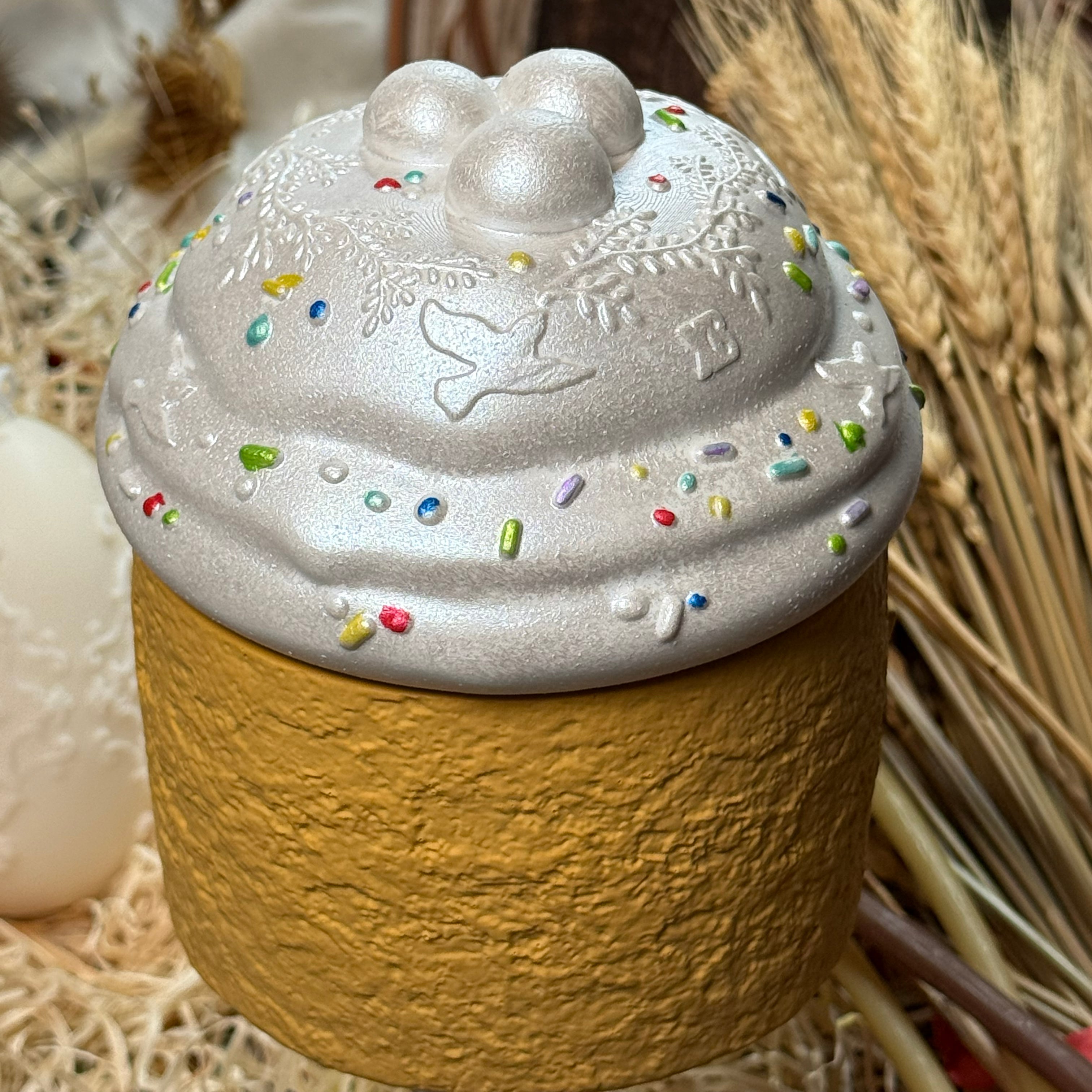 A detailed macro shot of the Easter Paska candle's lid, showcasing the intricate relief patterns of doves, floral branches, and colourful raised sprinkles on a pearl-white hand-painted finish.