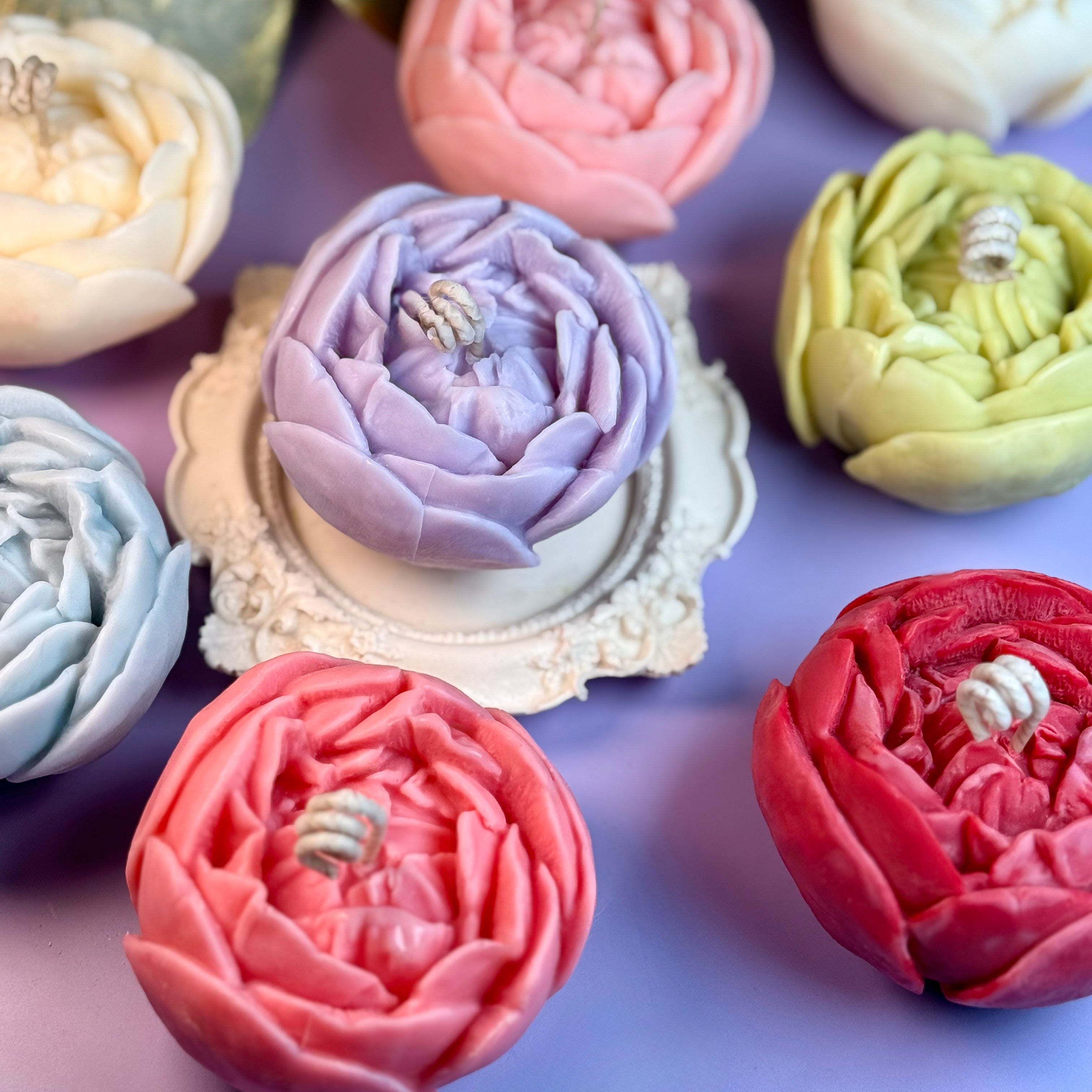 A collection of colourful artisan peony candles in pastel pink, purple, olive green, soft blue, yellow, red, and ivory white, arranged together on a purple backdrop.