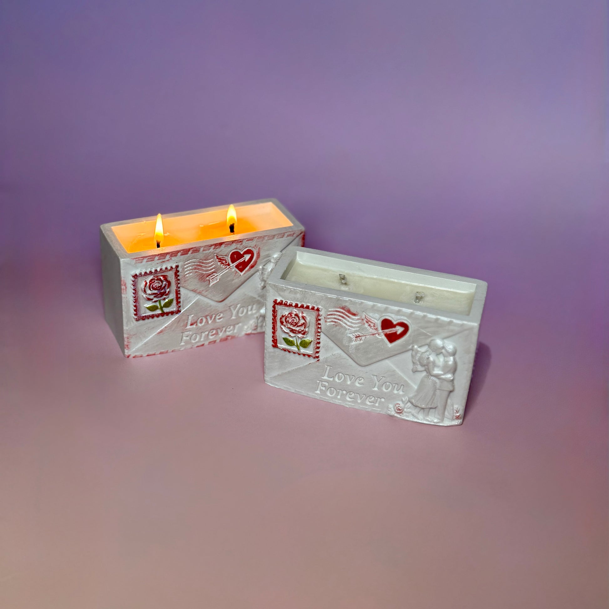 Two handcrafted love letter soy candles arranged on a pastel background, one unlit in the foreground showing the romantic relief carvings, and one burning softly behind it.