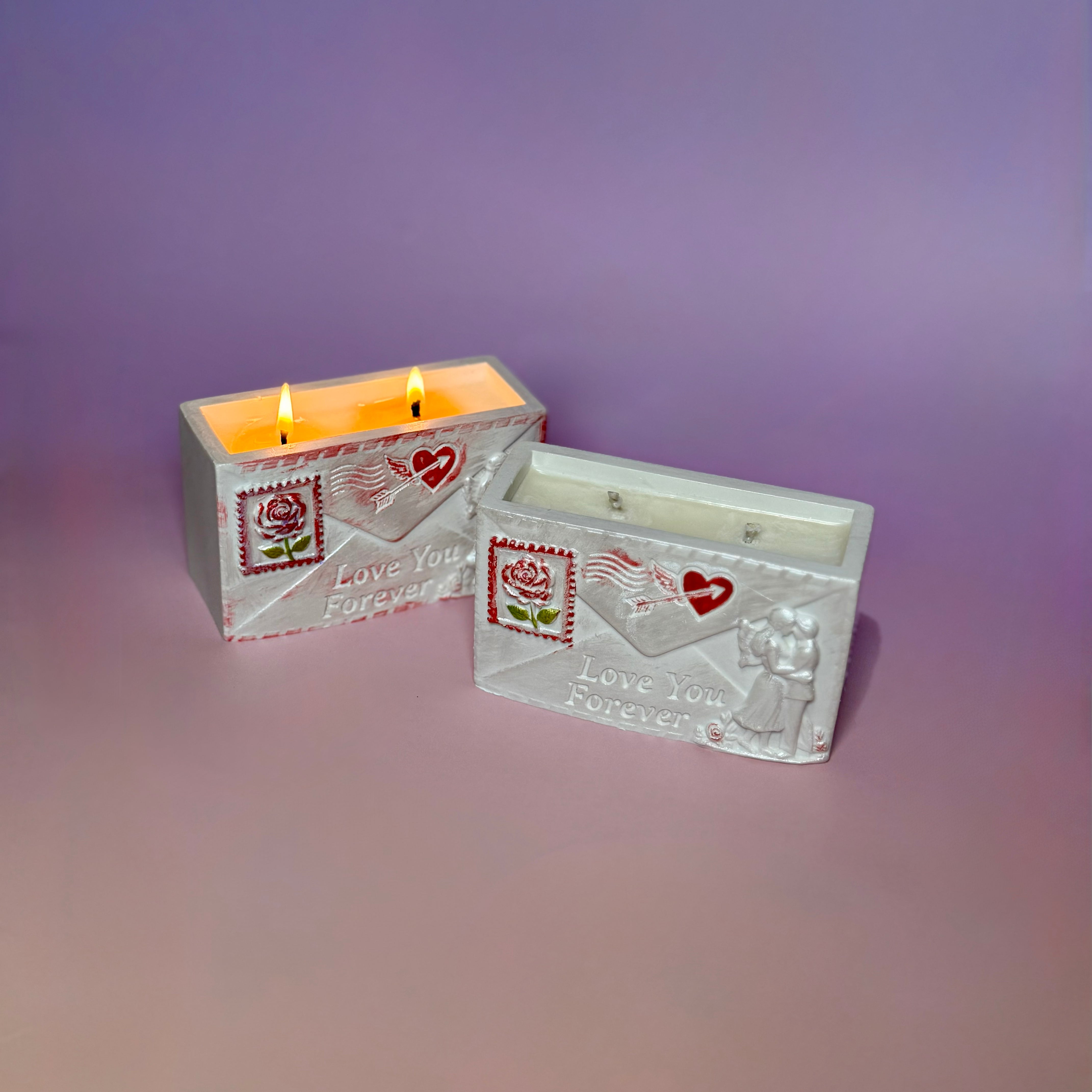 Two handcrafted love letter soy candles arranged on a pastel background, one unlit in the foreground showing the romantic relief carvings, and one burning softly behind it.