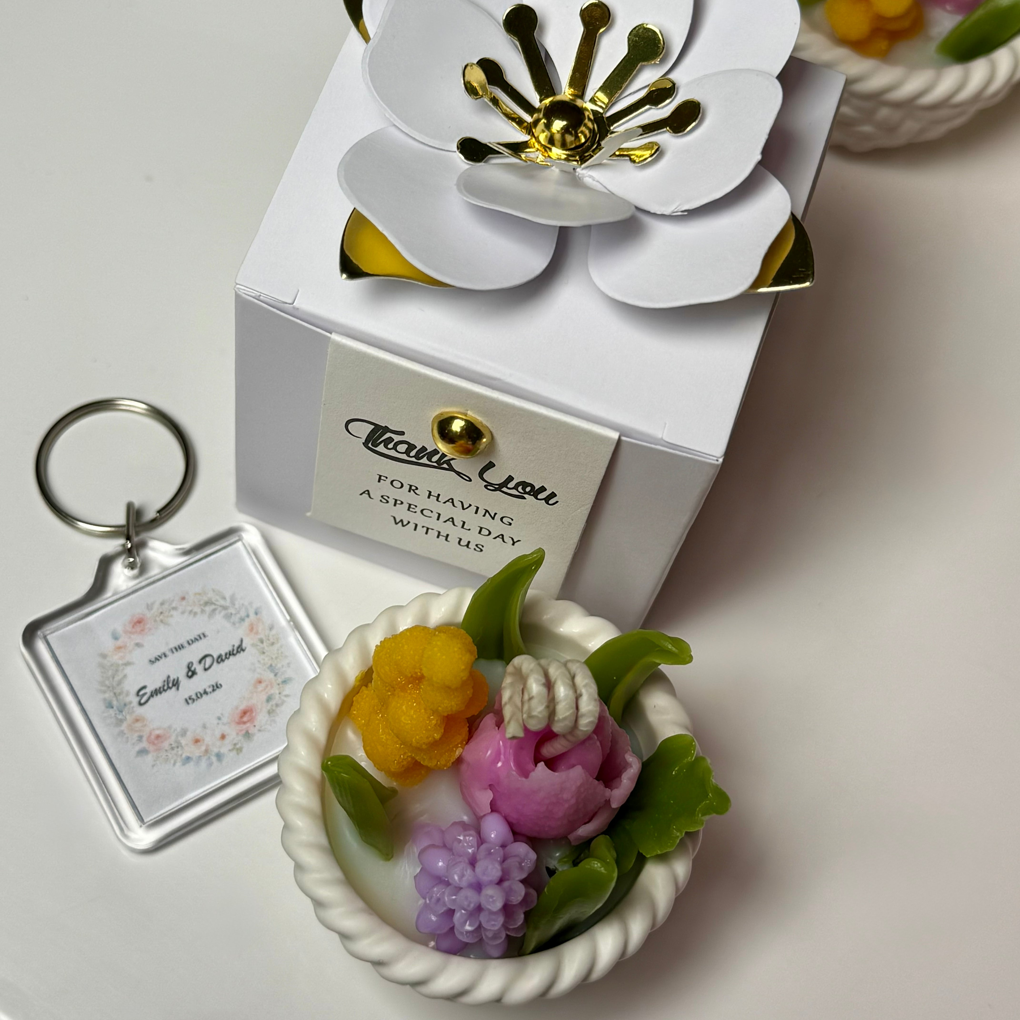 Top view of a personalised wedding favour set showing a square acrylic keychain printed with names and date, a white favour box with a luxury paper flower, and a colourful botanical basket candle.