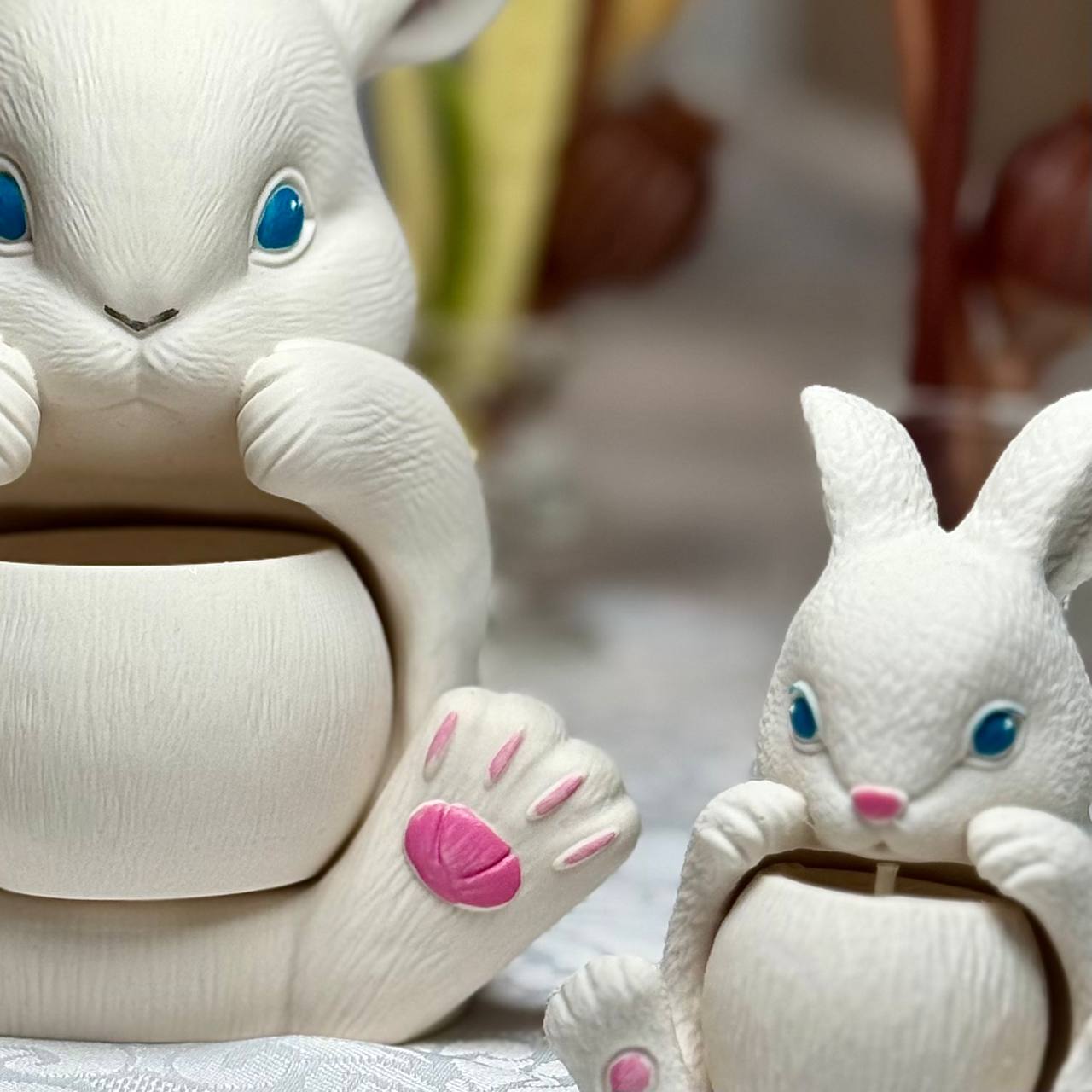 A close-up shot focusing on the hand-finished details and smooth white texture of the large and small Jesmonite bunny candle vessels on a white cloth.