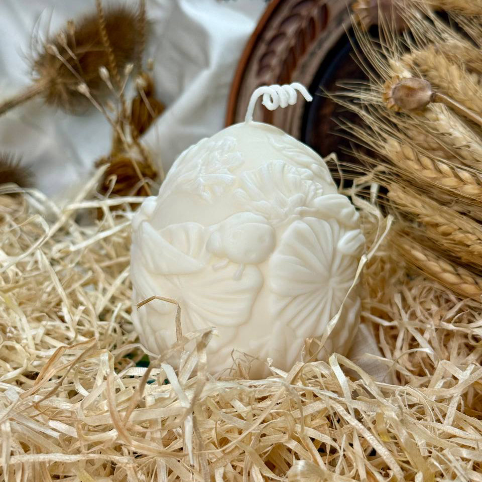Alternate angle of the large botanical relief egg candle, highlighting a sculpted ladybug on a flower leaf with dried wheat stalks and a wooden frame in the background.