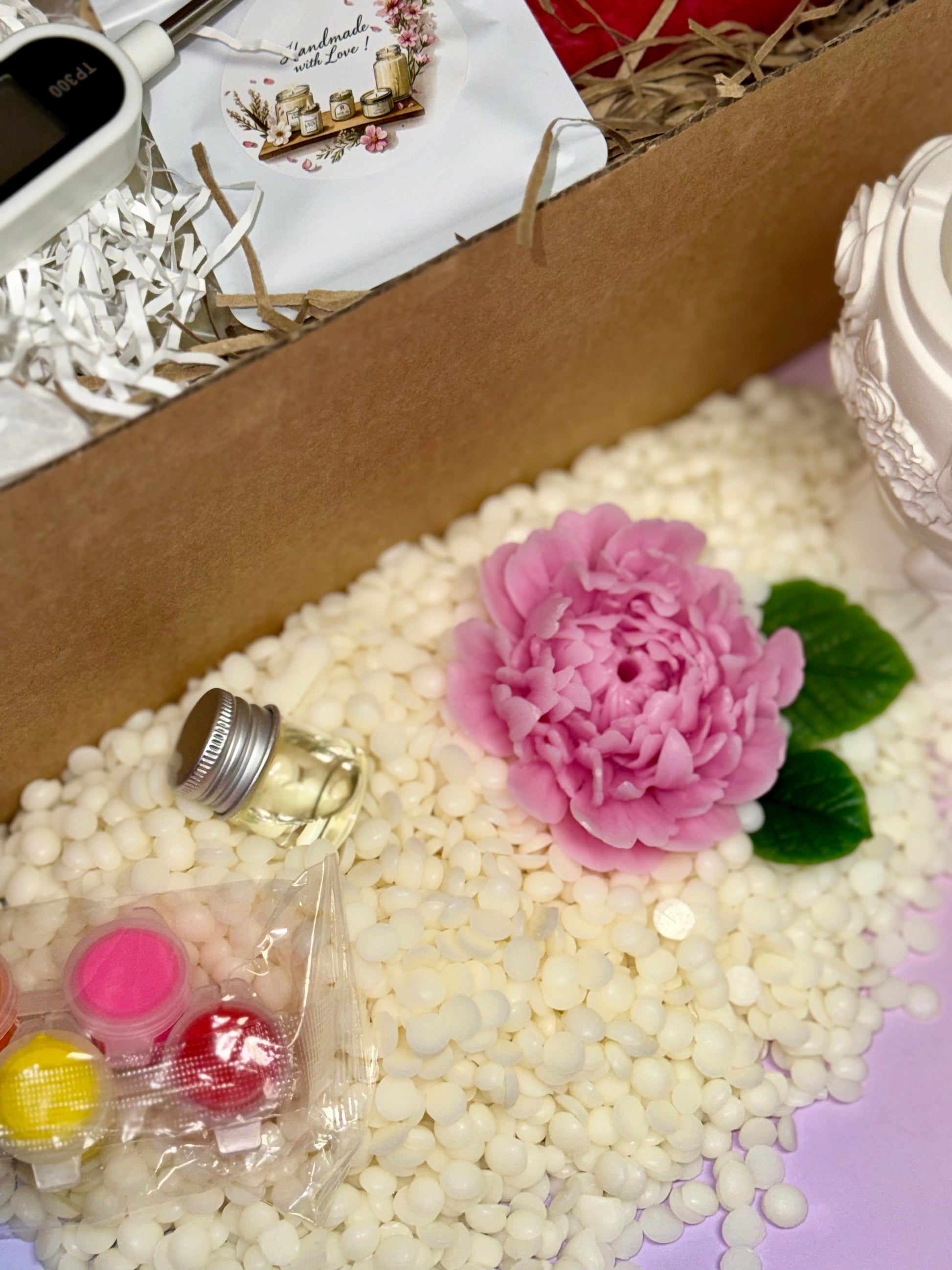 A close-up shot of the candle crafting ingredients, featuring a pile of creamy white soy wax pellets, a detailed pink beeswax peony flower, green leaves, colour pods, and a small glass vial of fragrance oil.