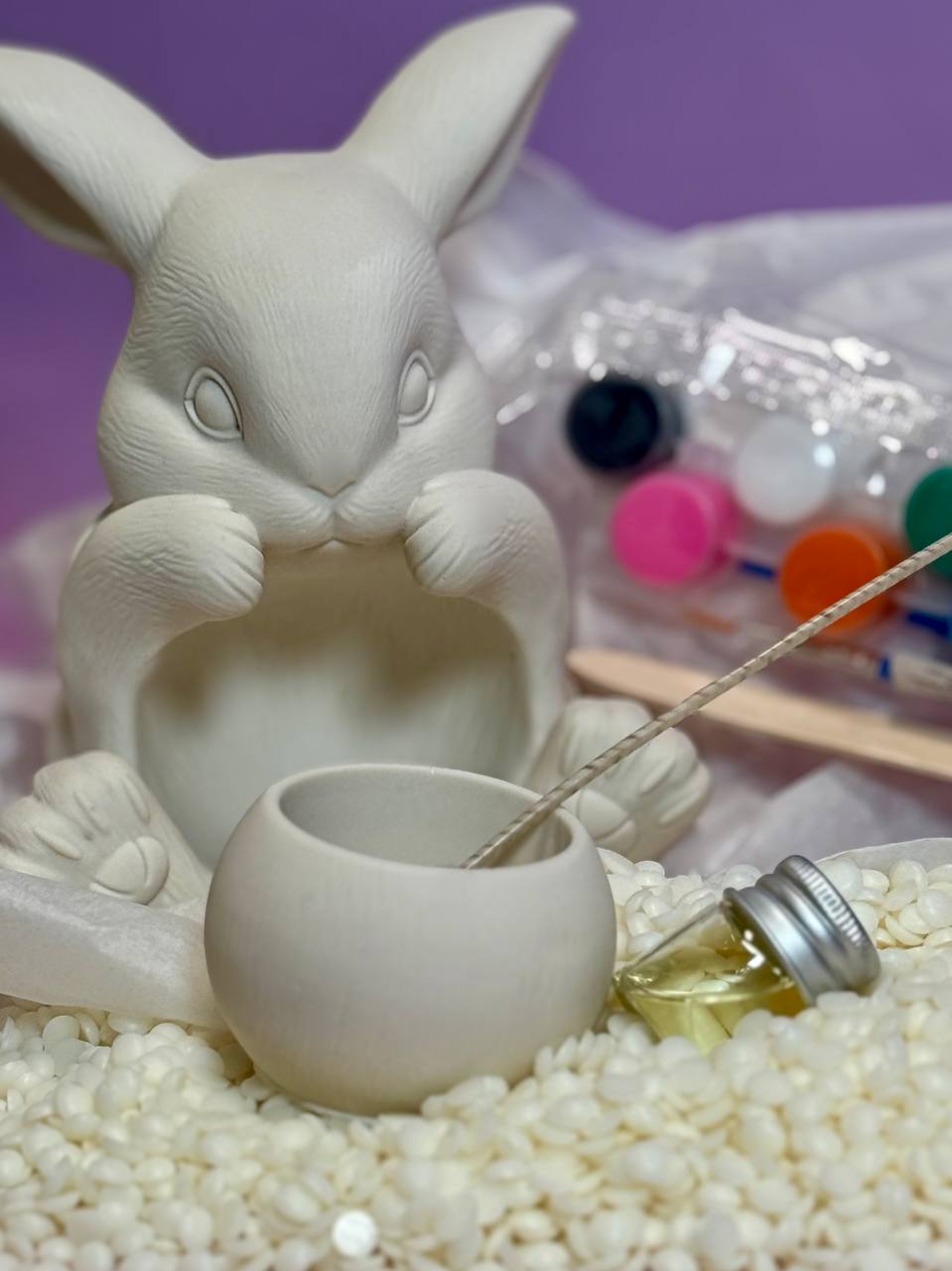 Complete components of a DIY Easter bunny candle making kit UK showing an unpainted Jesmonite bunny vessel, soy wax flakes, fragrance oil bottle, cotton wick, and acrylic paints with a brush.