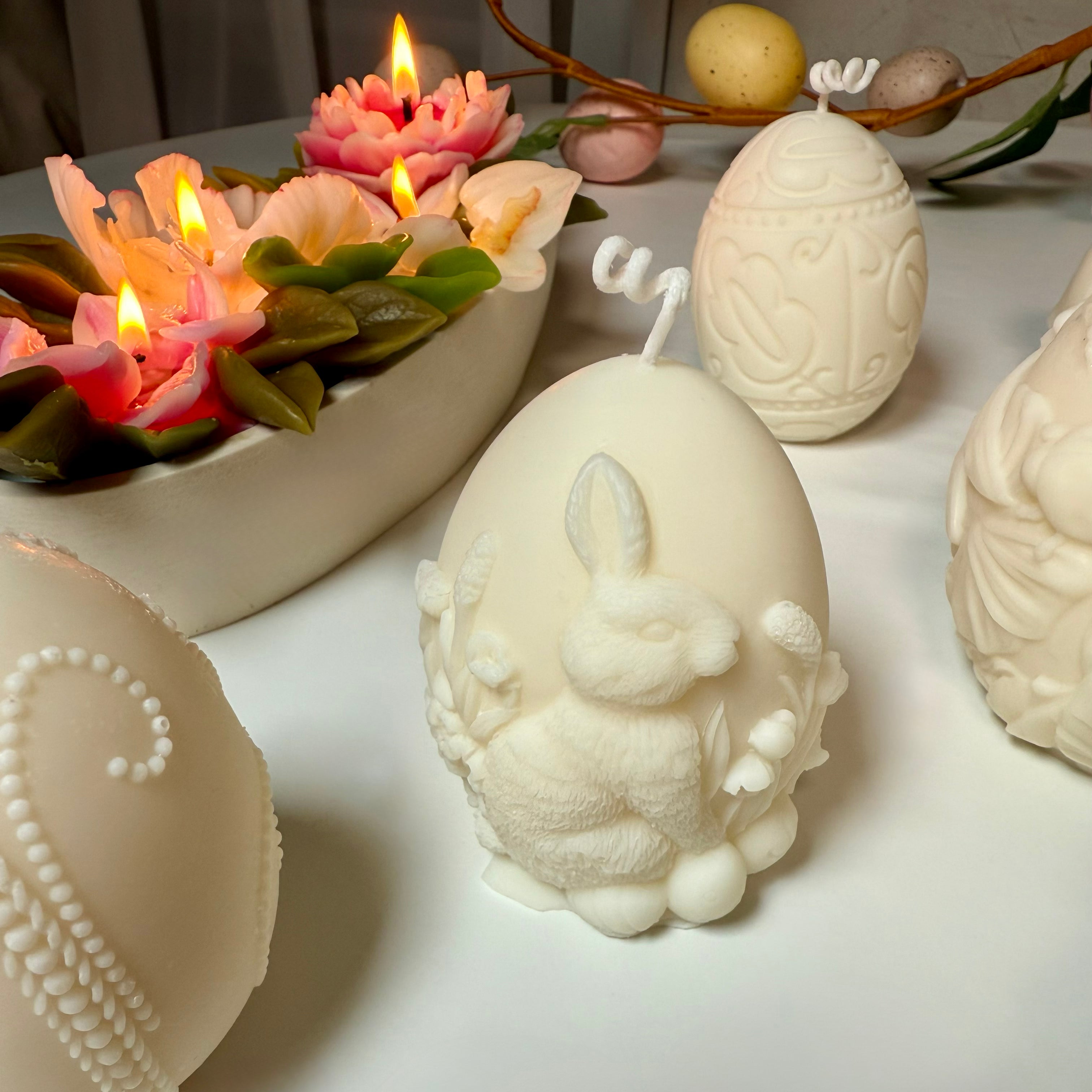 An artisan Easter table centrepiece display featuring the unburnt 3D bunny egg candle on a white tabletop alongside other decorative moulded shaped candles and burning floral candles.