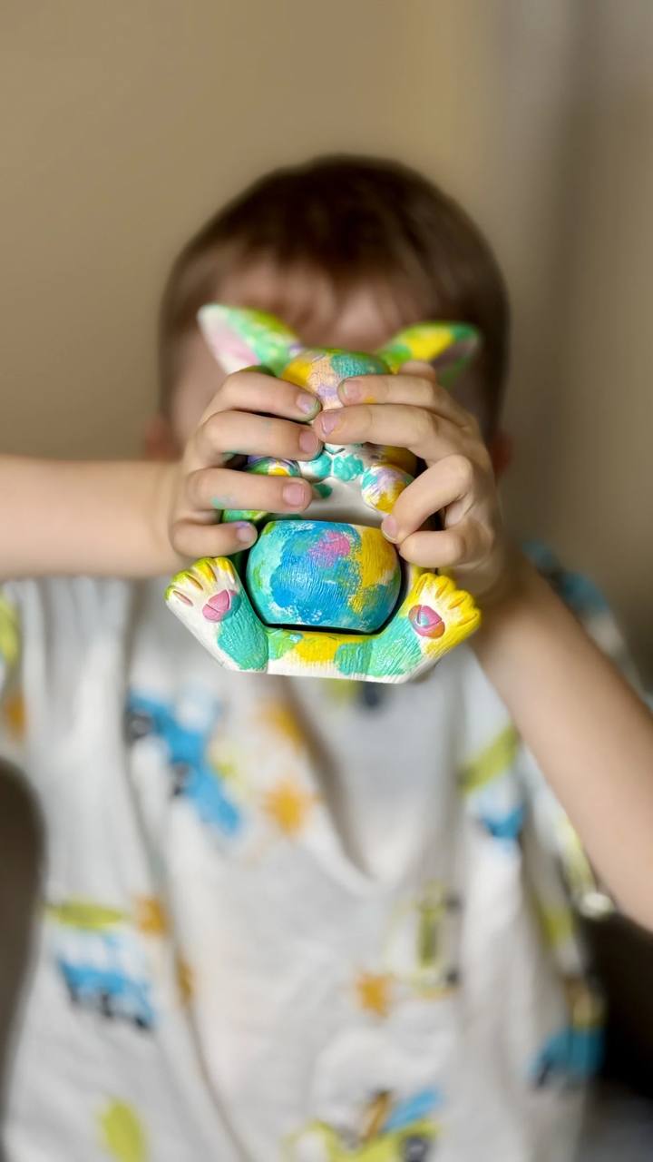 Two completed and hand-painted Jesmonite bunny candle vessels, large and small, with custom colourful designs and blue eyes, standing on a tablecloth next to wax flakes and dried floral stems, showing the results of an eco-friendly craft kit project.
