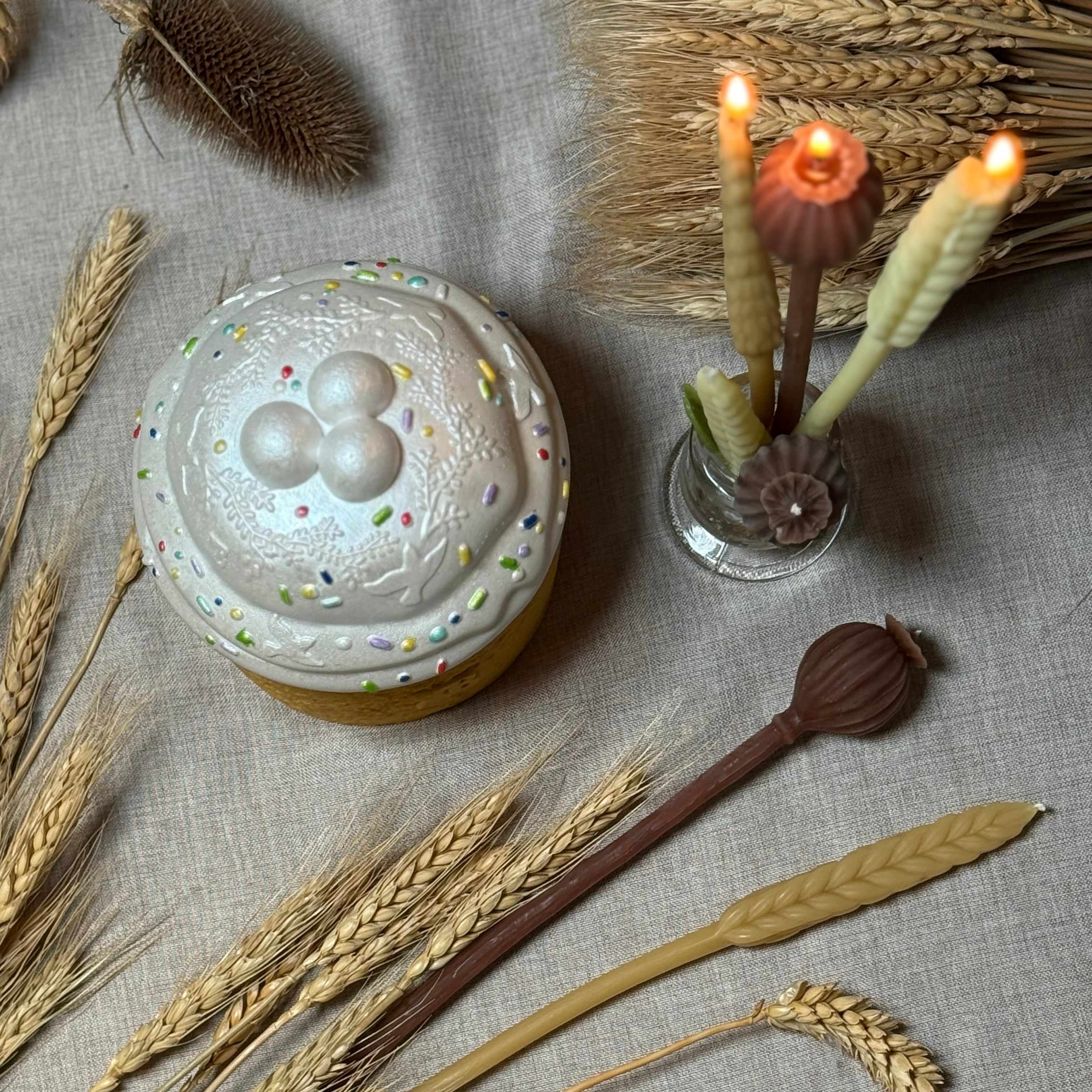 A flat lay composition of the pearl-topped Paska candle surrounded by natural dried wheat stalks and lit tapered beeswax candles shaped like wheat ears and poppy seed heads on linen fabric.
