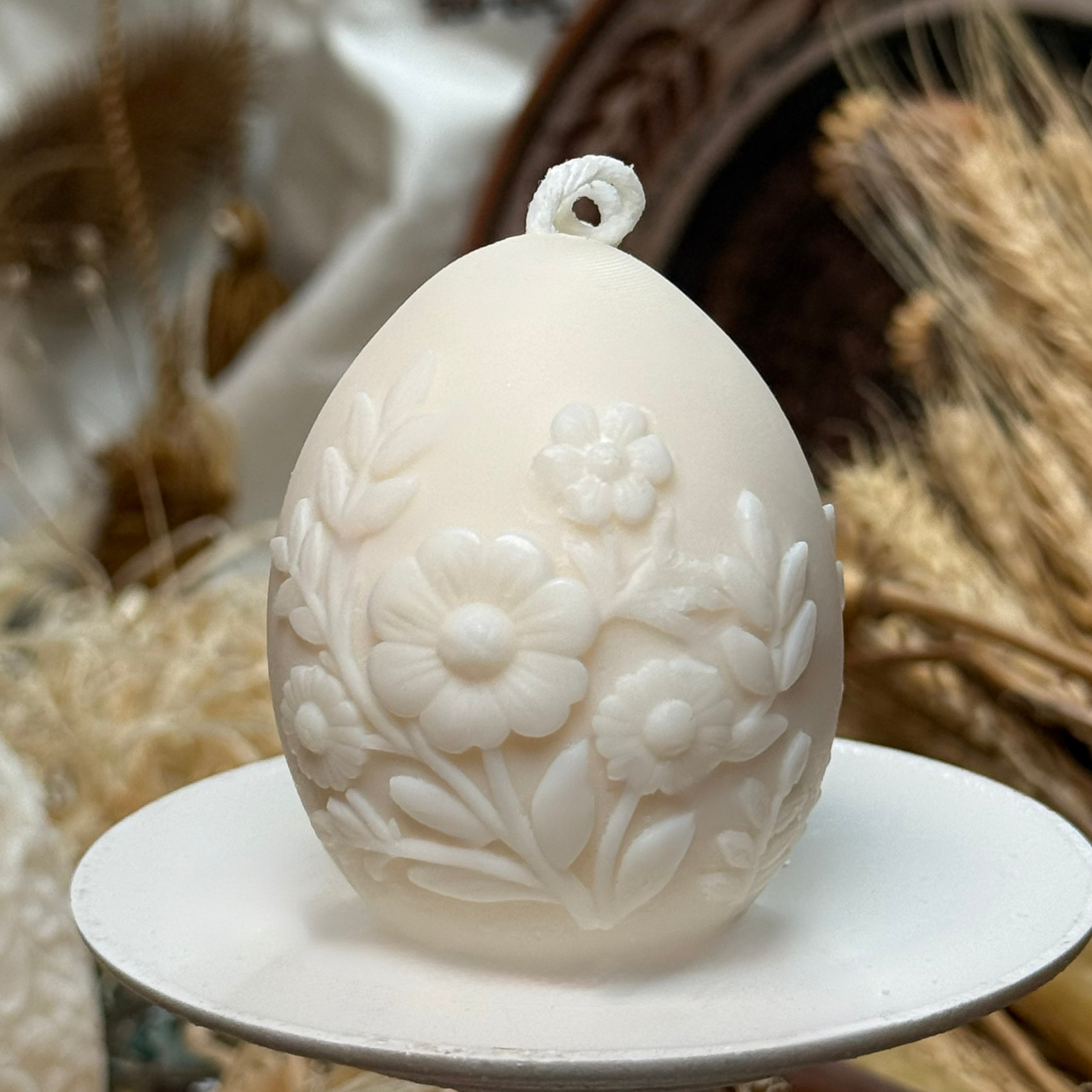 A front-facing view of the creamy white natural soy wax Easter egg candle on a white pedestal, showcasing the detailed 3D relief of daisies and spring meadow flowers wrapping around the smooth egg shape.