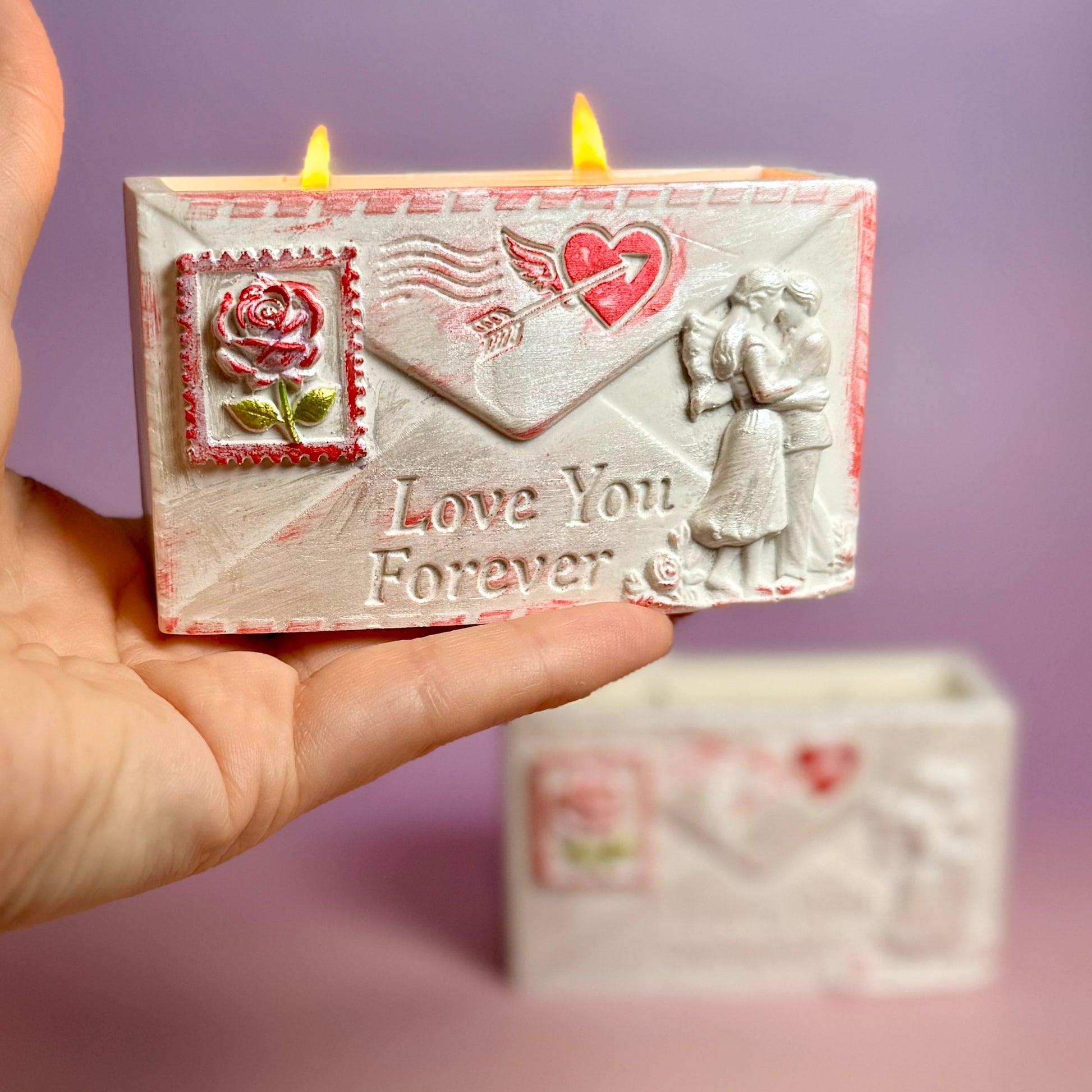 A hand holding the lit double-wick soy wax candle inside the handcrafted pearl-white love letter container, showcasing the red distressed edges and detailed couple carving.