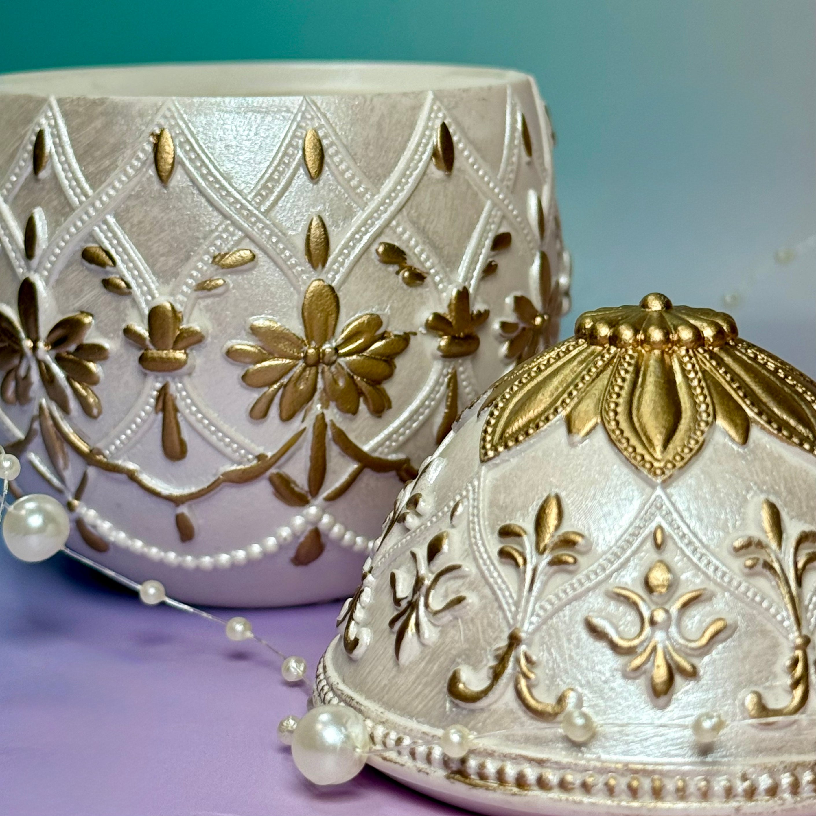Macro close up shot of the intricate relief patterns hand-painted with luxury gold detailing on a white pearlescent Easter egg candle lid and base.