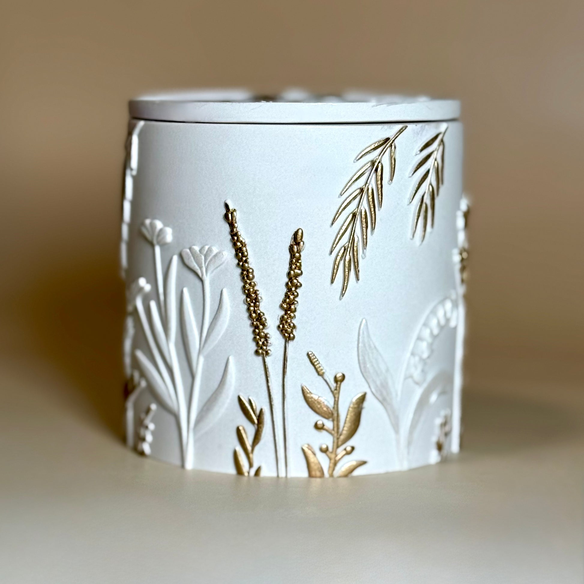 Front view of a closed botanical wildflower candle in a pearlescent white Jesmonite jar with a lid, featuring intricate relief patterns of meadow grass hand-painted with gold highlights.
