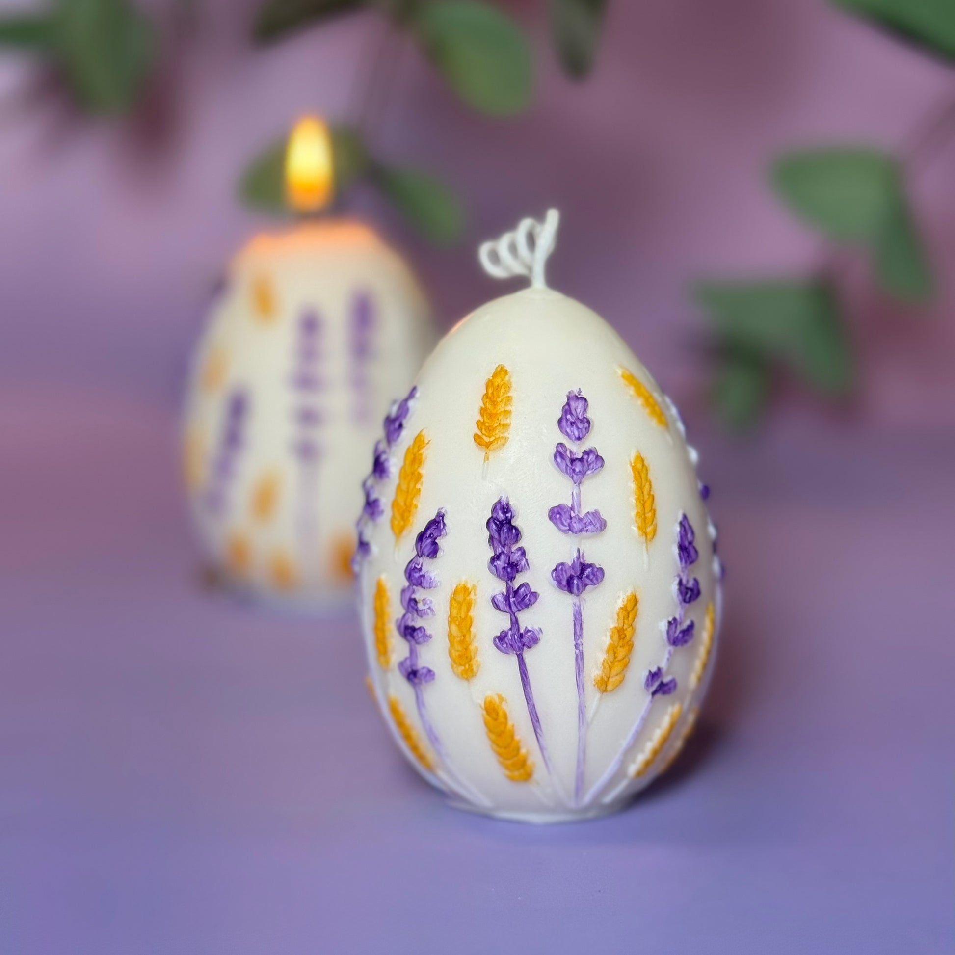 A close-up focus shot of a hand-painted soy wax Easter egg candle featuring golden yellow wheat stalks and purple lavender relief details against a soft pastel purple background.