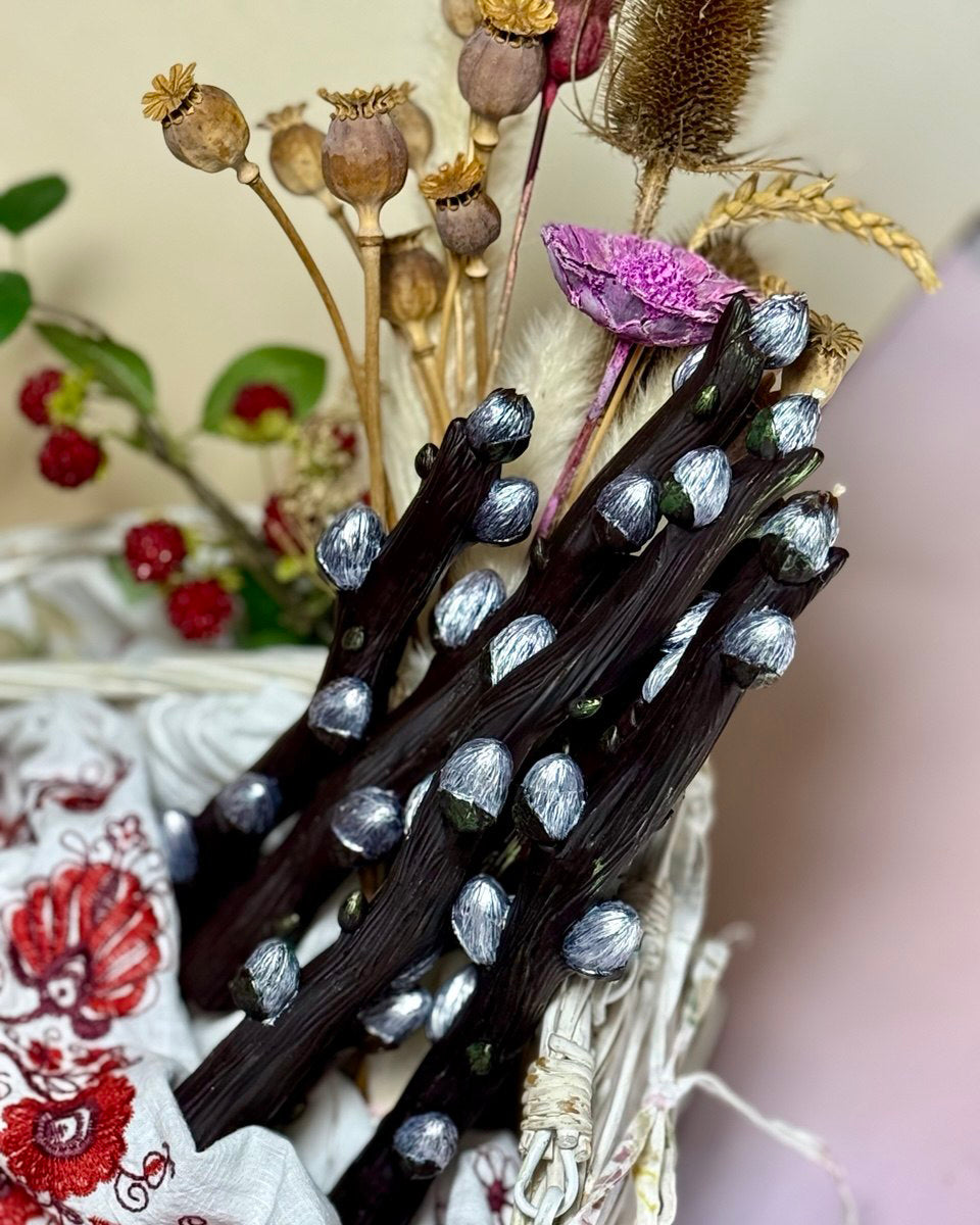 A bundle of detailed handcrafted beeswax pussy willow branch candles with realistic silver-painted buds gathered inside a white wicker basket with dried flowers.