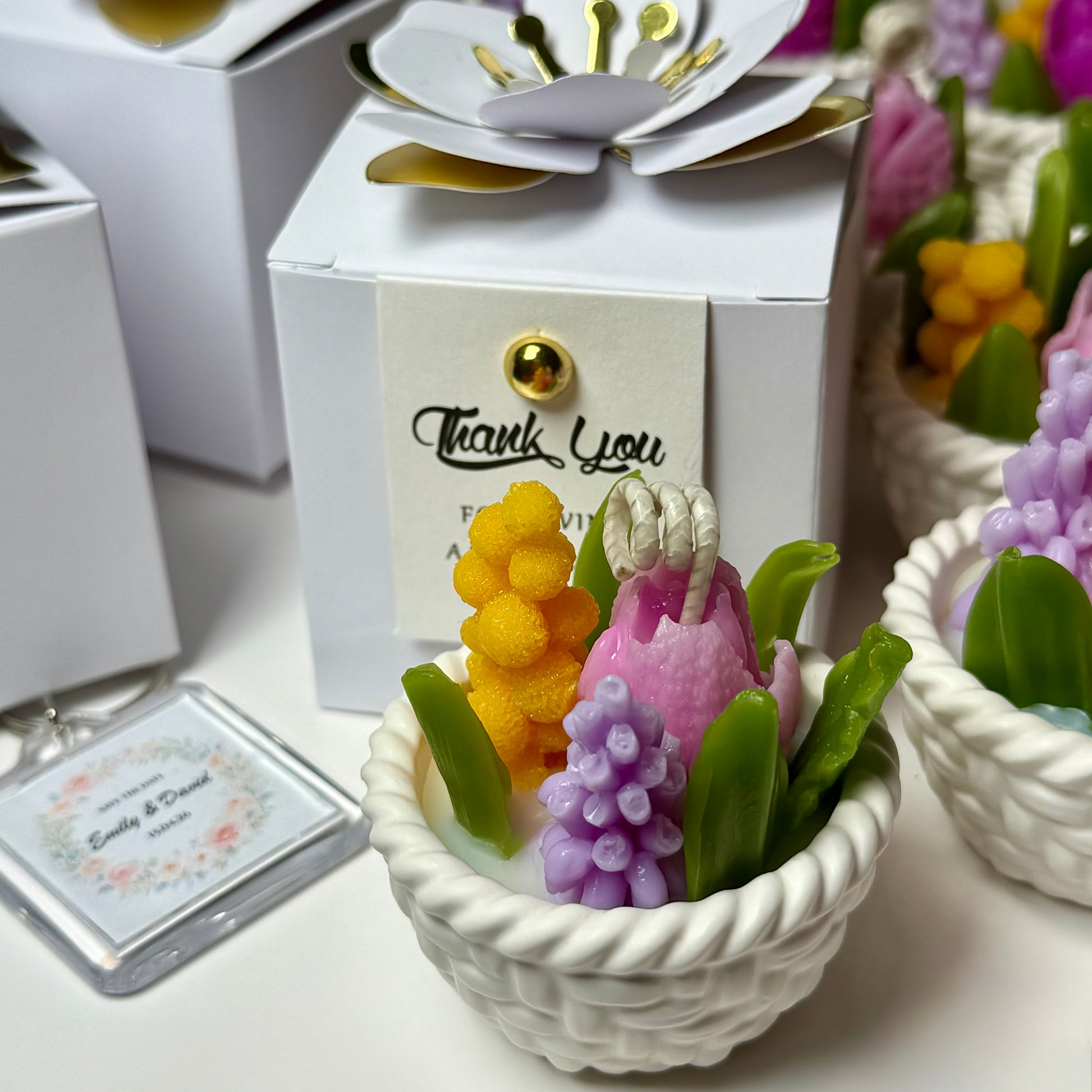 A close-up shot of a handcrafted botanical candle basket with purple, pink, and yellow beeswax flowers next to a white favour gift box and a personalised wedding keepsake keychain.