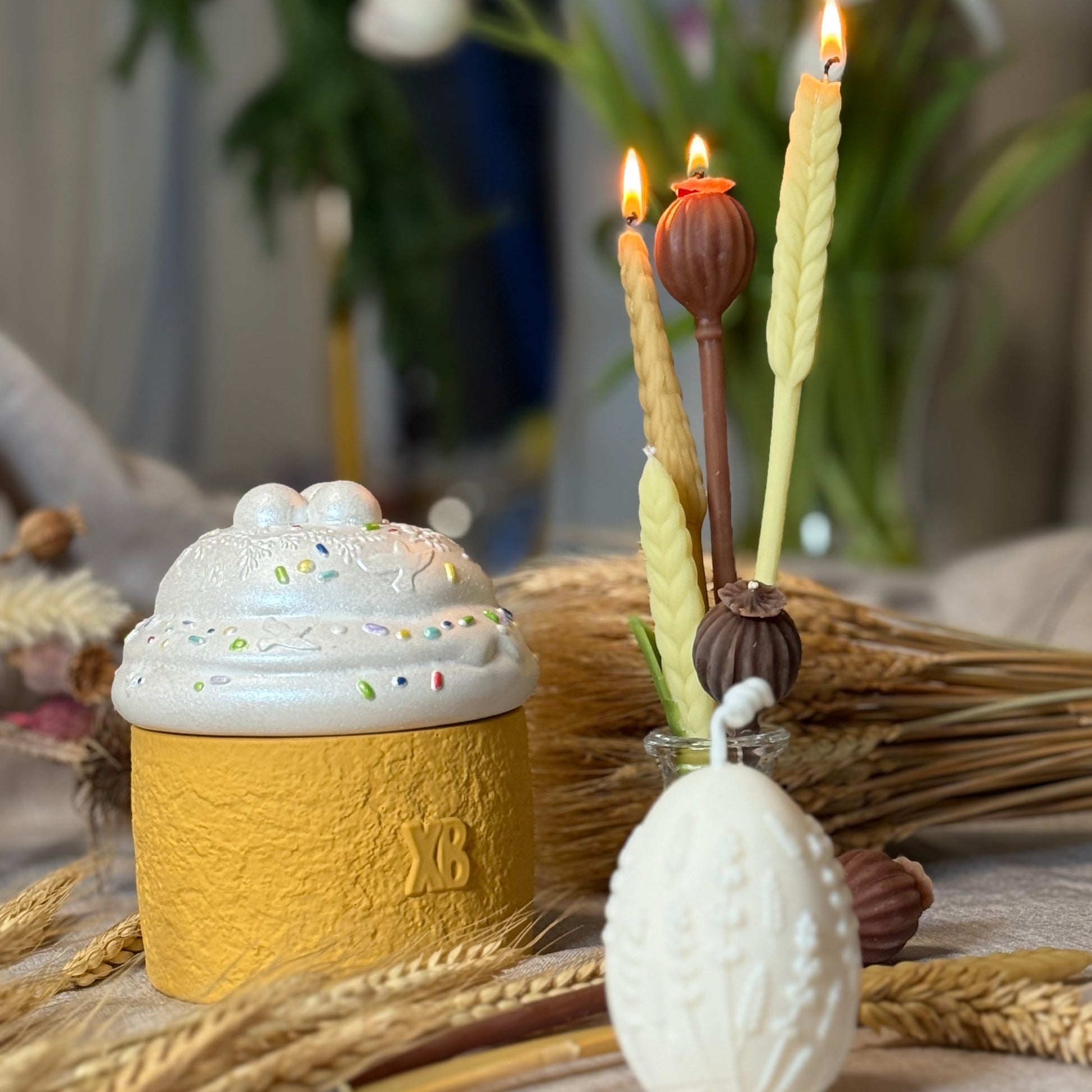 An elegant Easter tablescape display featuring the Paska bread candle with the traditional Cyrillic letters XB embossed on its textured yellow base, paired with burning beeswax wheat stalk and poppy pod candles.
