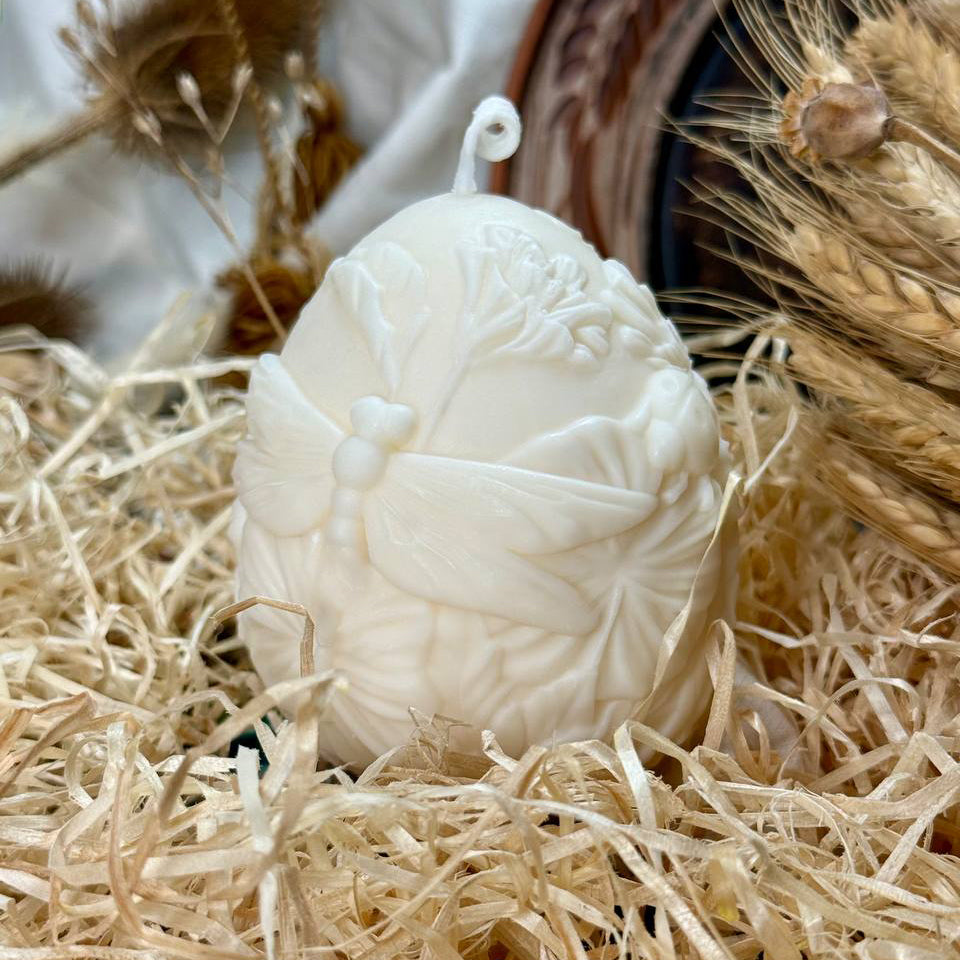 A large 250g creamy white soy wax Easter egg candle featuring a highly detailed 3D relief of a dragonfly among spring blooms, styled in a bed of rustic paper shred.