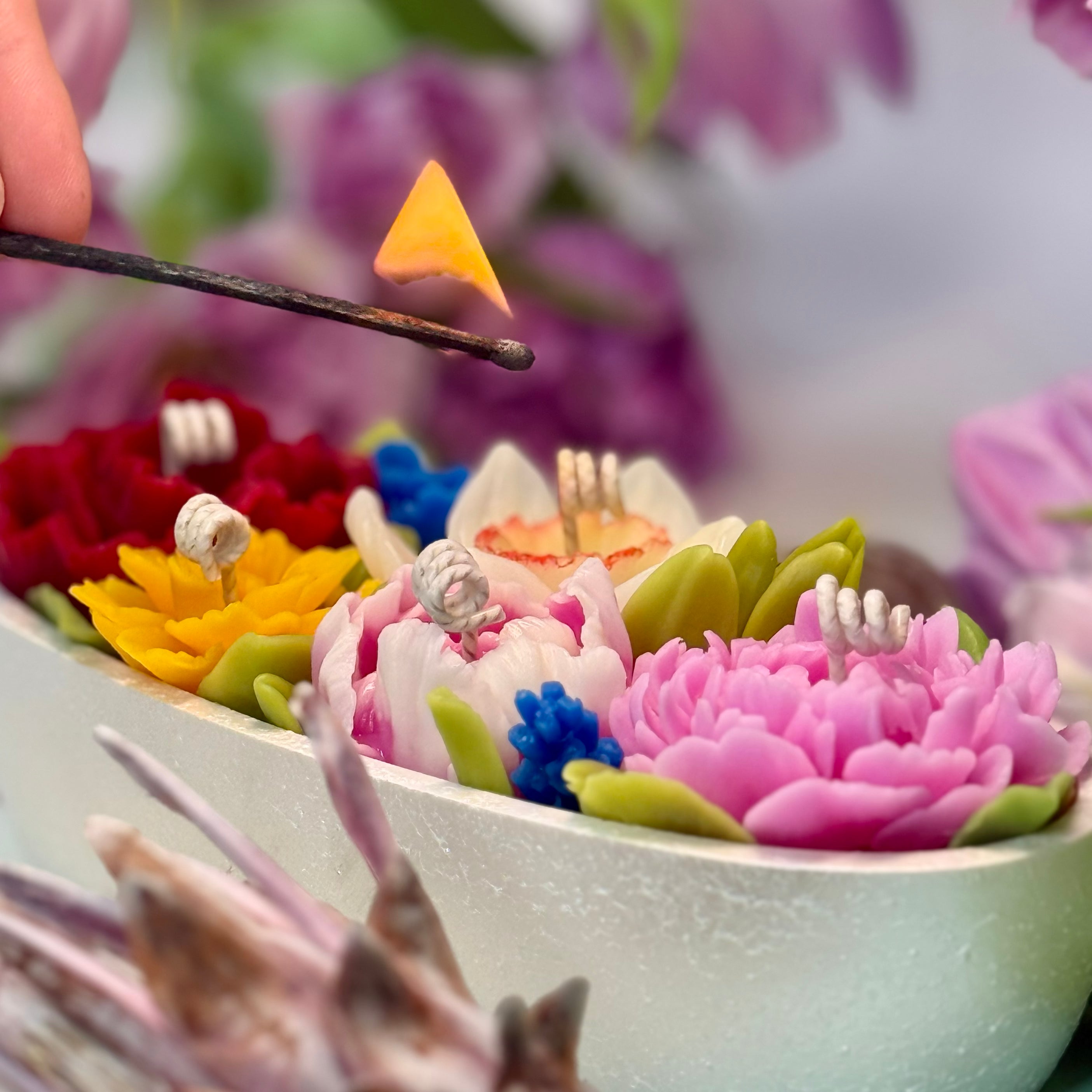 Macro photograph of a hand lighting the cotton wicks of intricately sculpted 3D beeswax flower candles embedded in a white Jesmonite vessel with gift box.